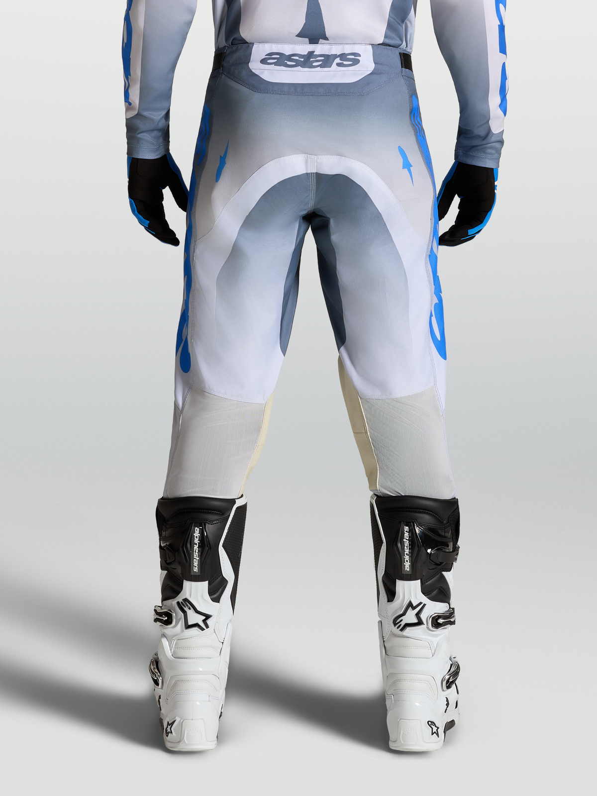 Alpinestars Pantaloni Racer Amber, pantaloni da motocross grigi e blu indossati con stivali bianchi, vista posteriore, design ergonomico pre-curvato per off-road