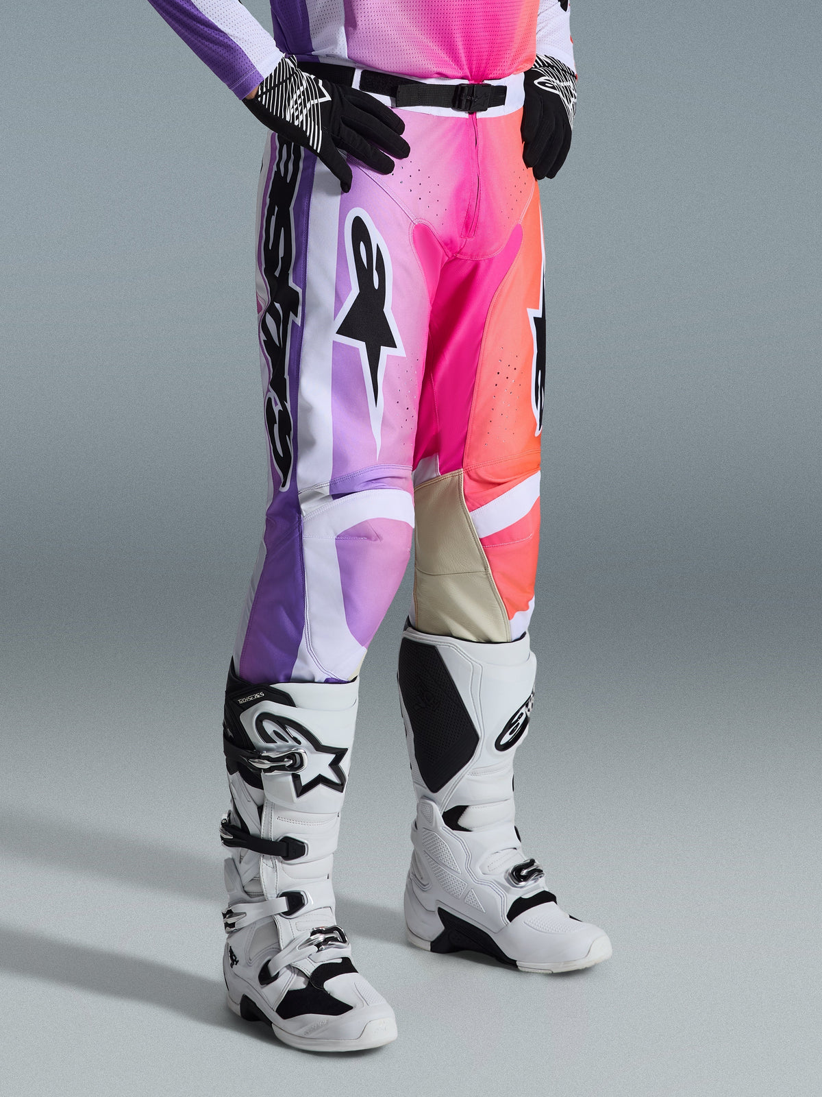 Alpinestars 2026 Racer Air Portl Pants, pantaloni da motocross, grafica multicolore con sfumature viola, rosa e arancione, indossati con stivali da motocross bianchi e neri e guanti neri, design race fit e pannelli interni alle ginocchia in pelle beige