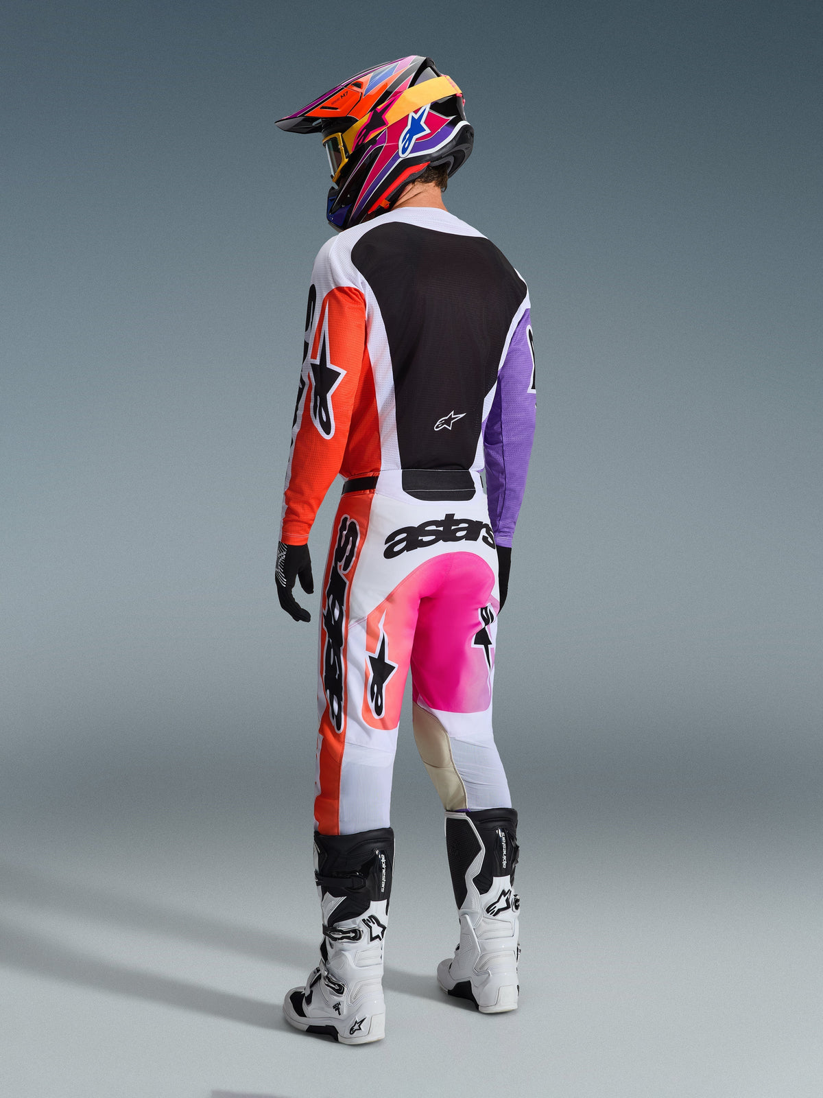 Pilota di motocross visto da dietro con i pantaloni Alpinestars 2026 Racer Air Portl in Multicolor, caratterizzati da pannelli bianchi, arancioni e viola con accenti rosa neon e loghi neri, abbinati a maglia coordinata, casco colorato e stivali bianchi e neri.