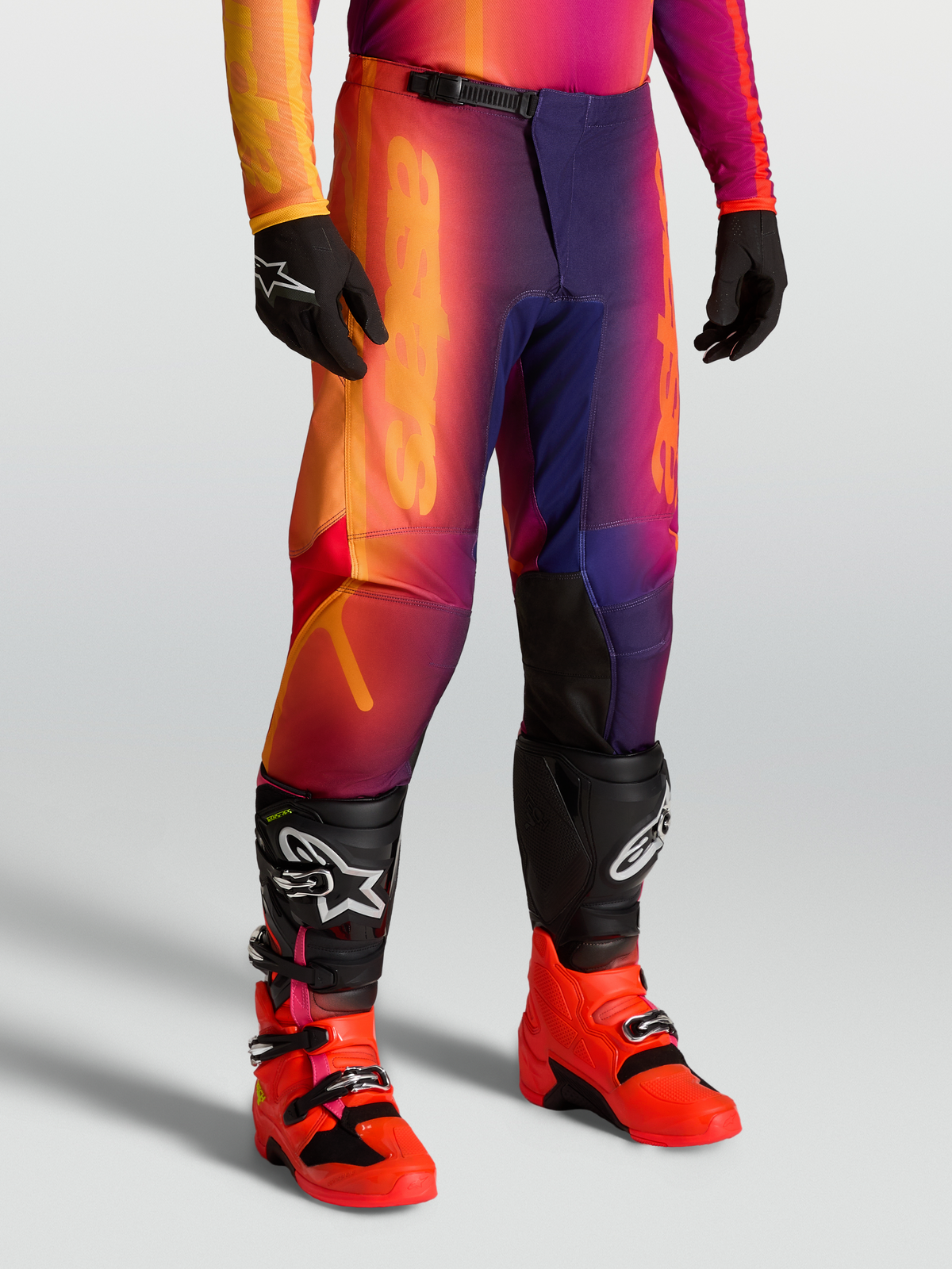 Alpinestars Pantaloni Fluid Kepler, pantaloni motocross sport-fit viola e arancione, con gambe pre-curvate e ginocchia rinforzate, indossati con stivali Tech serie.