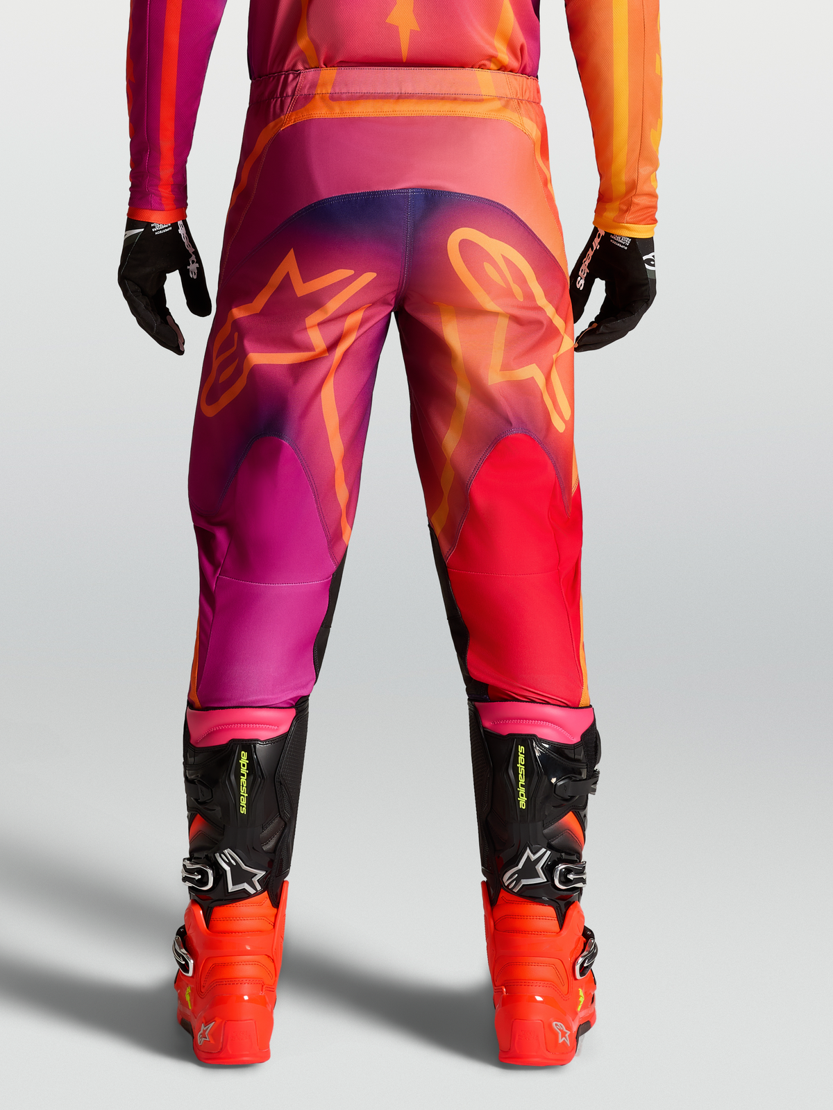 Alpinestars Pantaloni Fluid Kepler, pantaloni motocross da uomo, viola e arancione, con grafiche a stella posteriori e stivali neri.