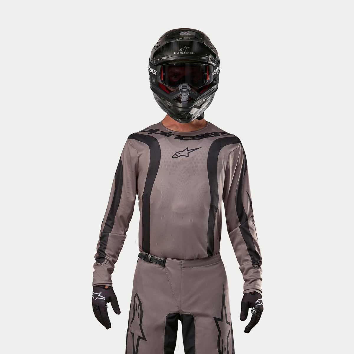 Pilota di motocross che indossa la maglia Alpinestars Fluid Lurv 2024 in Mud Black, un tessuto grigio-marrone terroso con strisce grafiche nere e logo a stella sul petto, abbinata a un casco nero, occhiali rossi e guanti neri per un look da corsa off-road coordinato