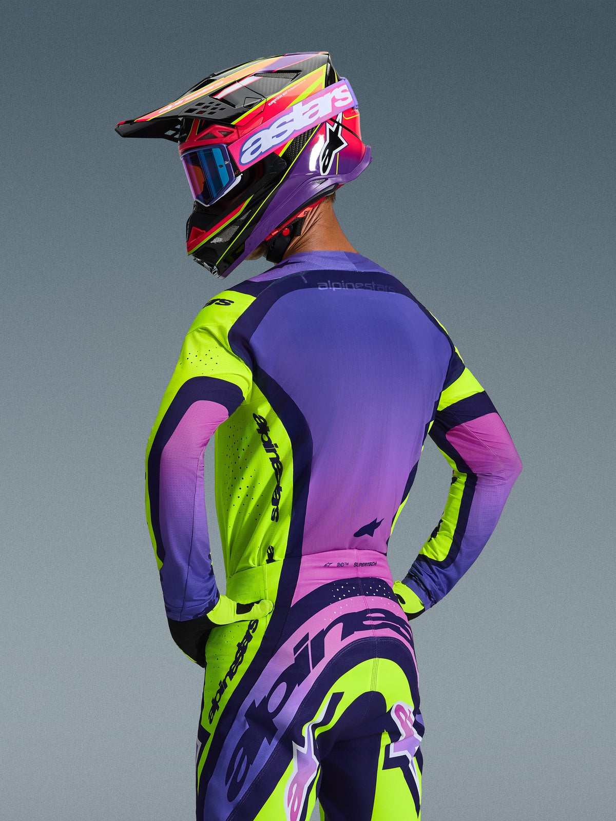 Pilota di motocross visto da dietro che indossa la maglia Alpinestars Supertech Vista 2026 e i pantaloni coordinati, colorazione Yellow Purple Black con sfumature vivaci, casco da corsa coordinato con occhiali blu e guanti gialli, kit professionale da fuoristrada con vestibilità atletica e loghi integrati.
