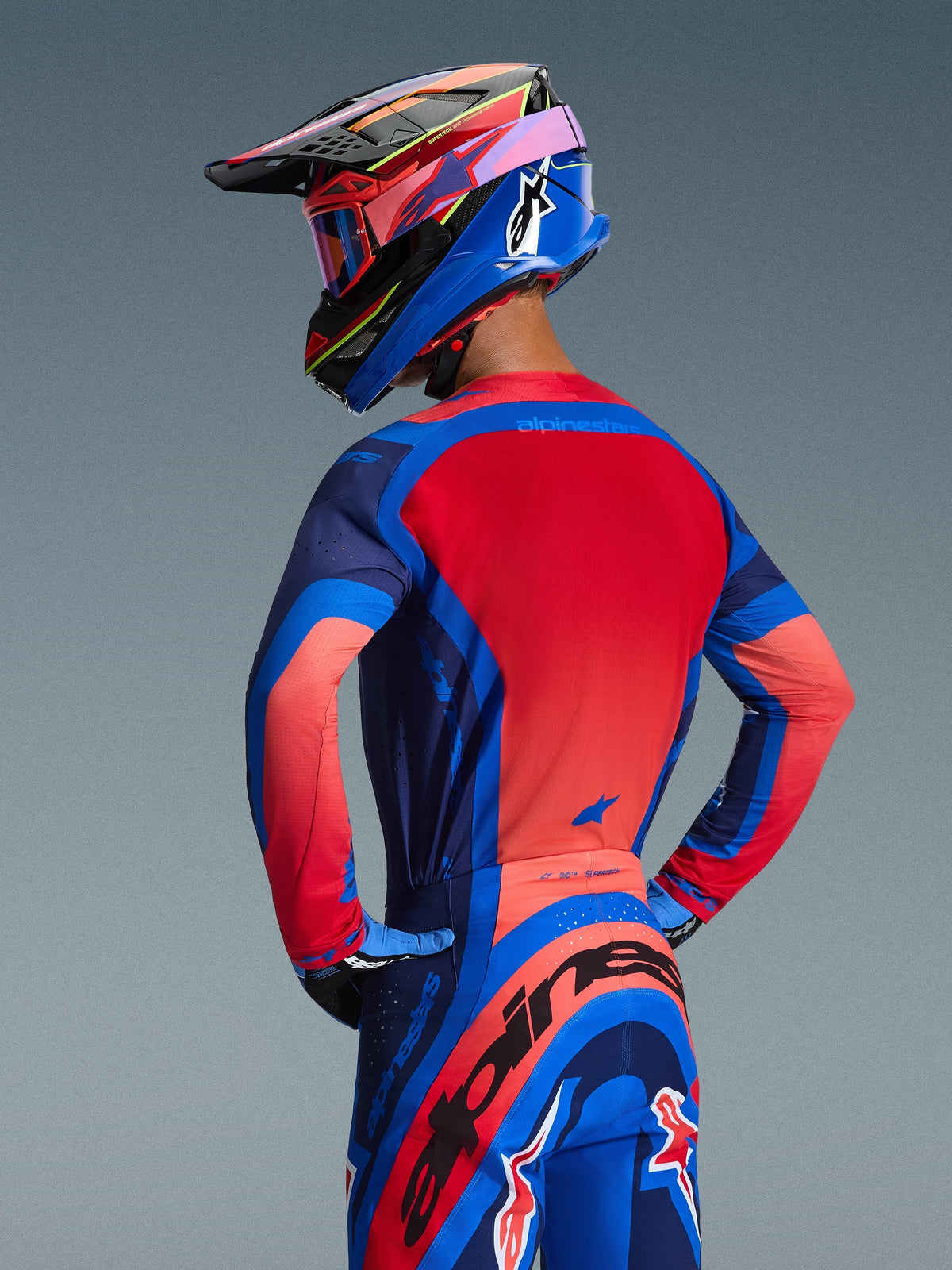 Vista posteriore di un pilota di motocross con maglia Alpinestars Supertech Vista 2026 nei colori Dark Navy, Hot Coral e Cobalt, caratterizzata da un vivace pannello posteriore rosso e costruzione senza cuciture, abbinata a pantaloni e casco coordinati per un look da gara completo.