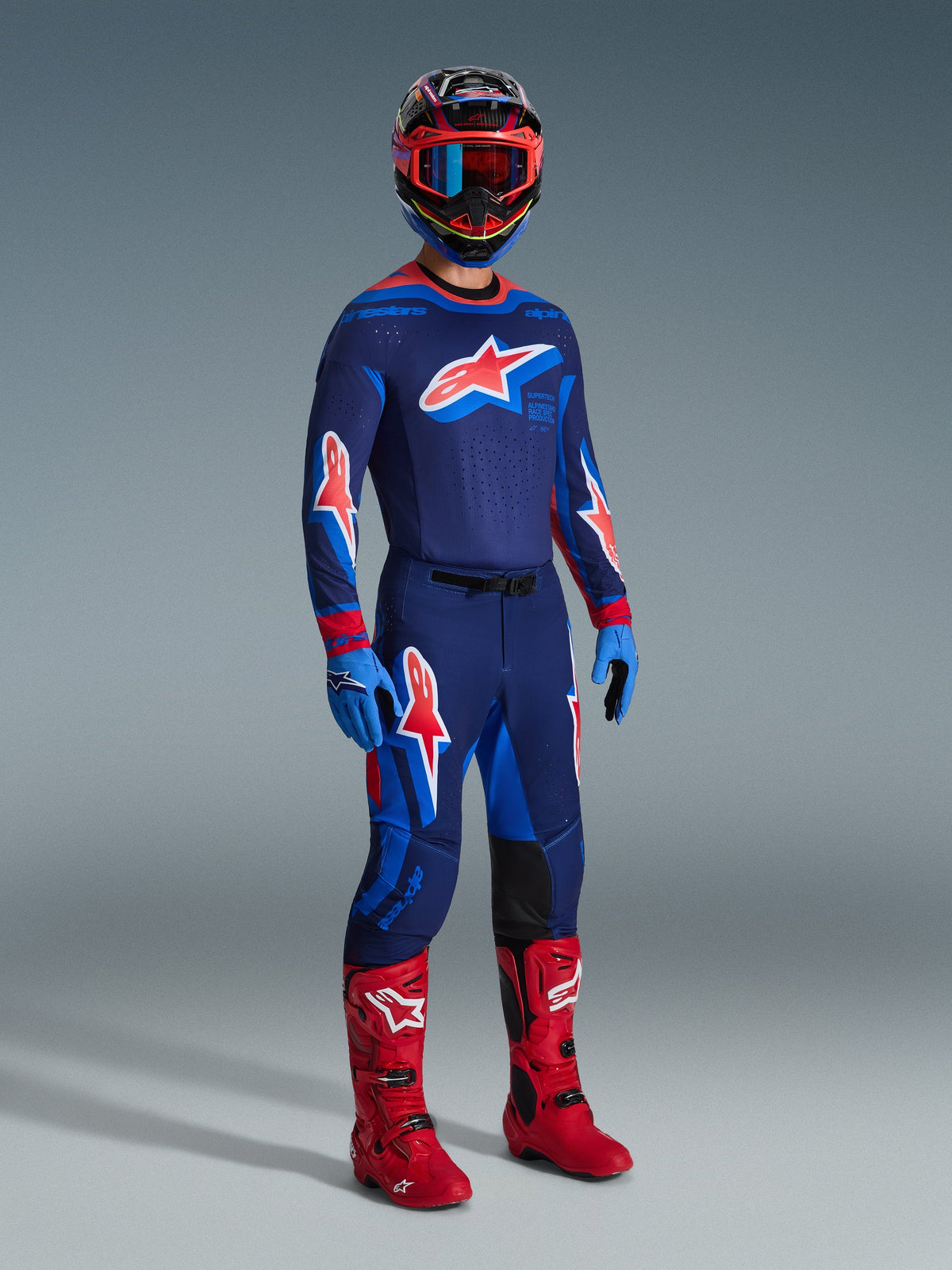 Pilota di motocross con equipaggiamento completo da gara, maglia Alpinestars 2026 Supertech Vista in Dark Navy, Hot Coral e Cobalt, pantaloni coordinati con grandi loghi a stella, stivali da motocross rosso acceso, guanti blu e casco multicolore con occhiali rossi, set professionale per fuoristrada