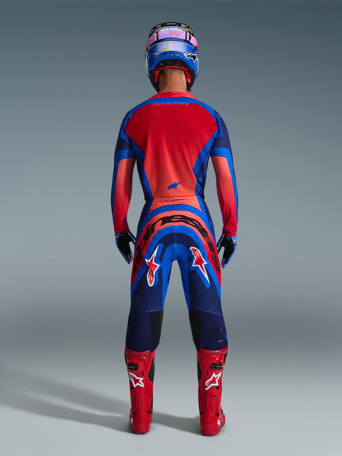 Pilota di motocross visto di spalle che indossa la maglia Alpinestars Supertech Vista 2026 in Dark Navy, Hot Coral e Cobalt, completo coordinato con pantaloni, stivali da gara rossi e casco blu, abbigliamento professionale per gare off-road.