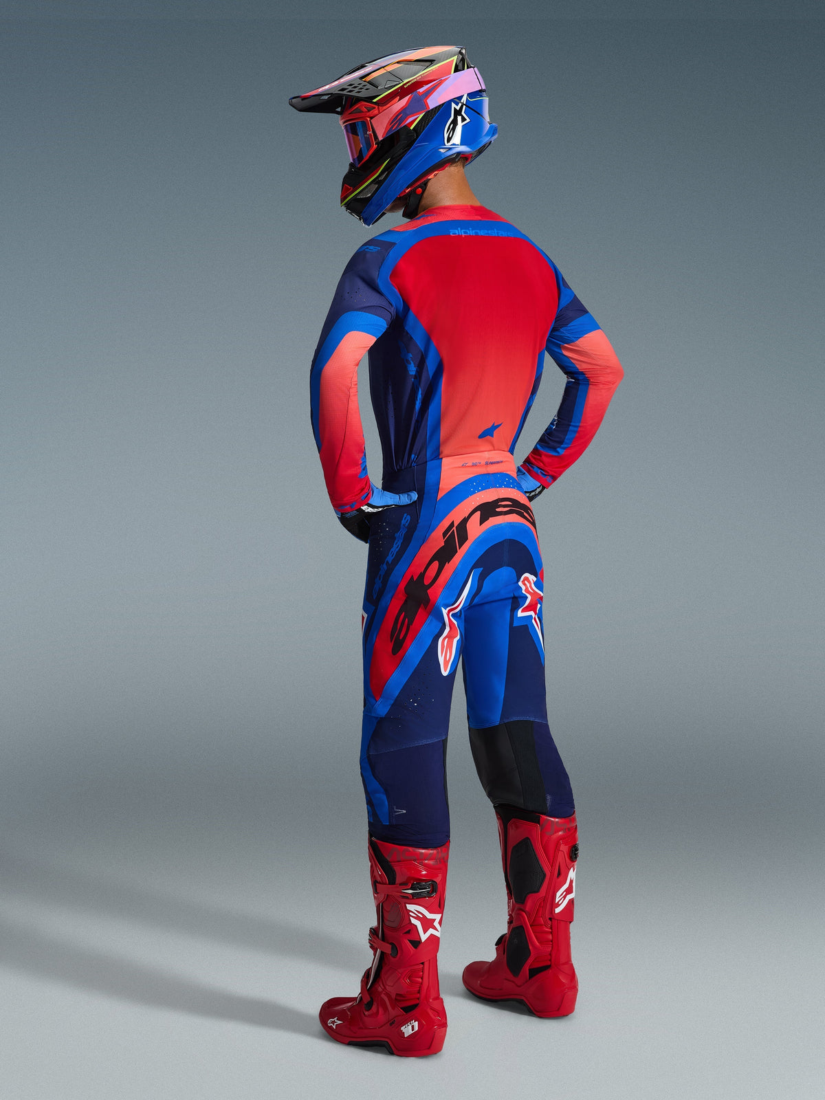 Pilota di motocross visto di spalle che indossa la maglia Alpinestars 2026 Supertech Vista in Dark Navy, Hot Coral e Cobalt, abbinata a pantaloni da motocross coordinati, stivali rossi e un casco colorato, abbigliamento professionale da gara coordinato