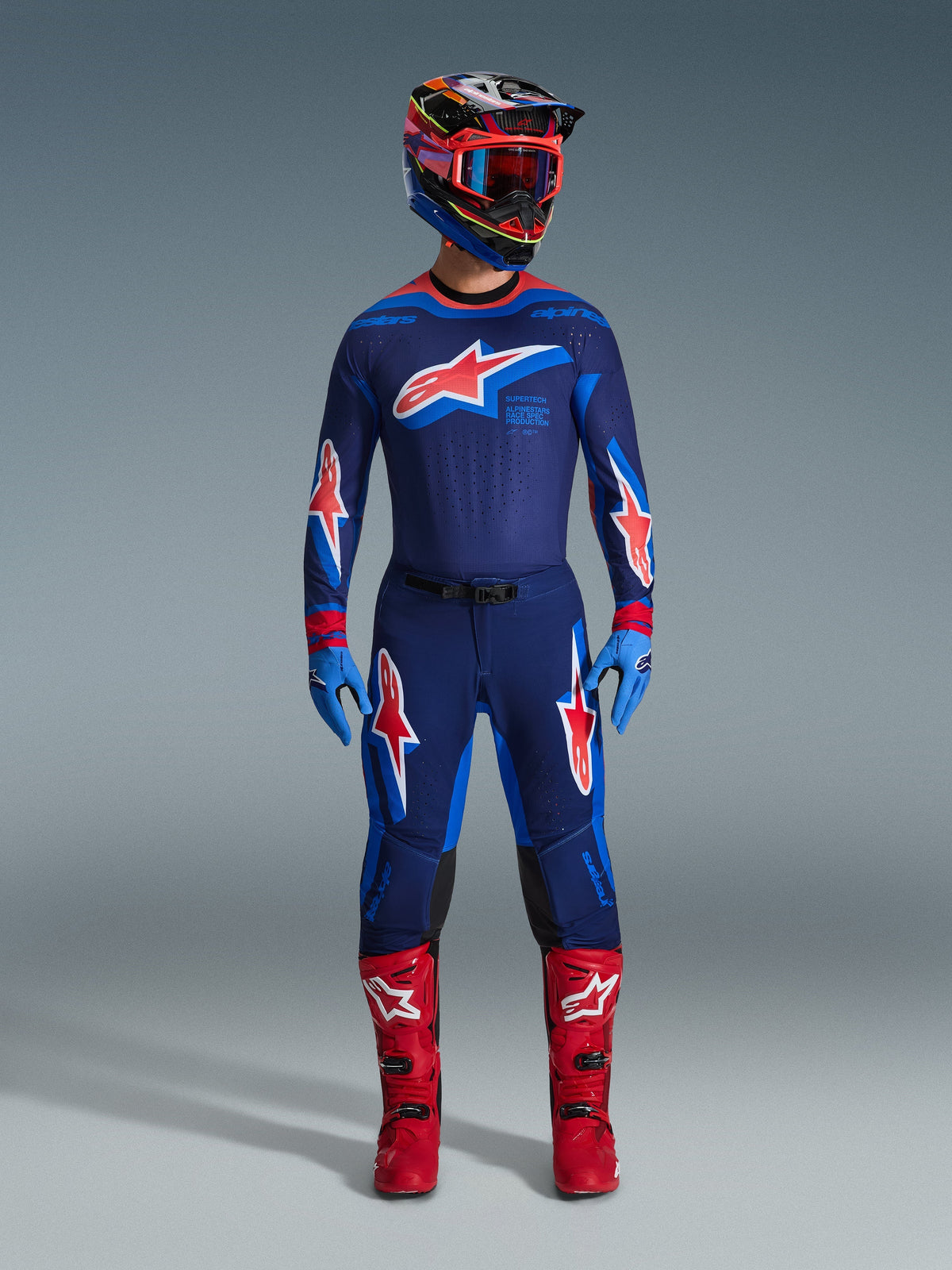 Pilota di motocross che indossa la maglia Alpinestars Supertech Vista 2026 in Blu Navy, Hot Coral e Cobalto, abbinata a pantaloni da gara coordinati, stivali rosso acceso e casco coordinato con occhiali rossi, set professionale completo per prestazioni fuoristrada.