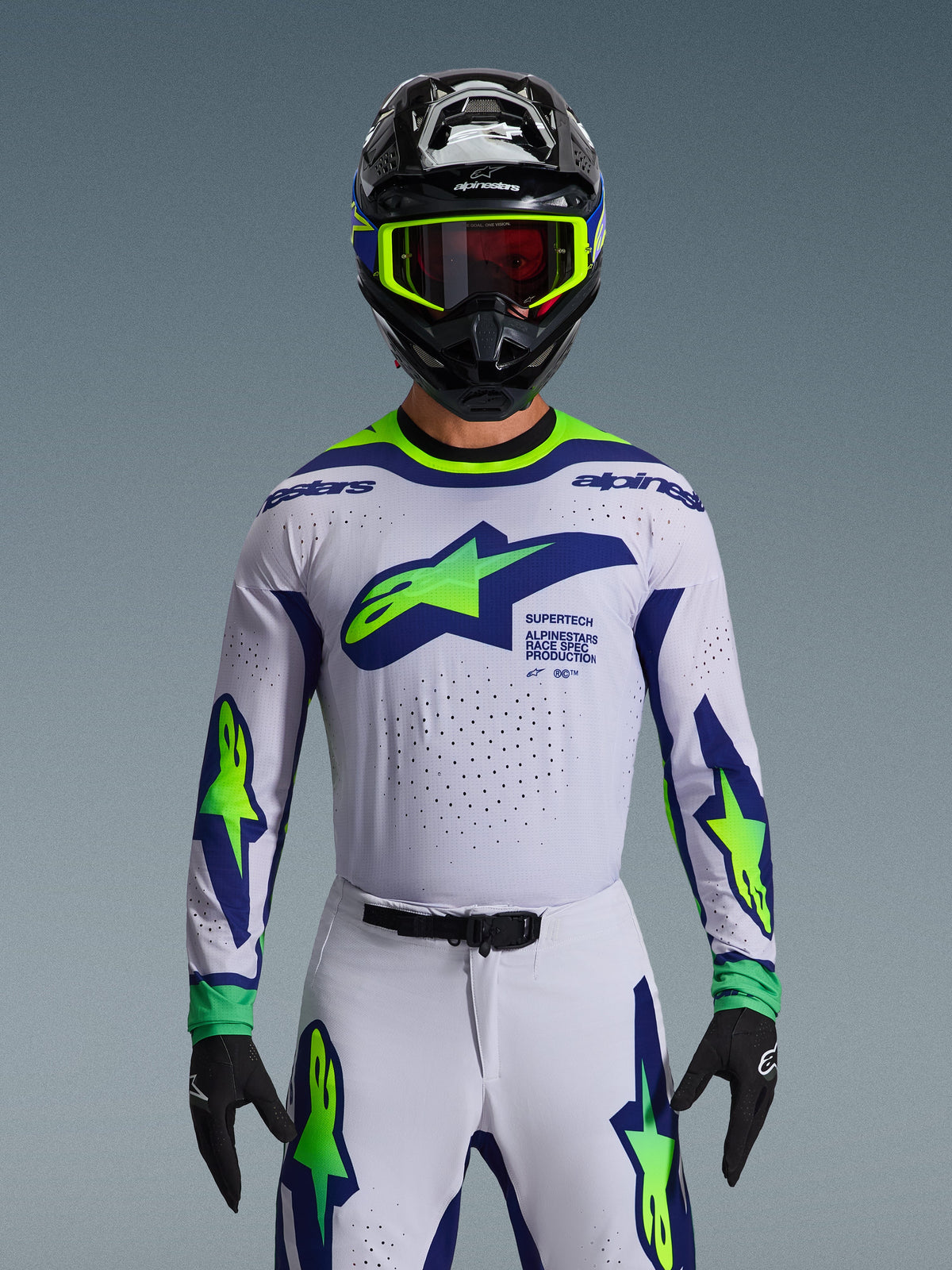 Alpinestars 2026 Supertech Vista Jersey, maglia da motocross, grigio chiaro, giallo fluo e blu, indossata da un pilota con pantaloni coordinati e casco nero, con un grande logo sul petto e perforazioni laser per la ventilazione, vestibilità atletica di livello professionale