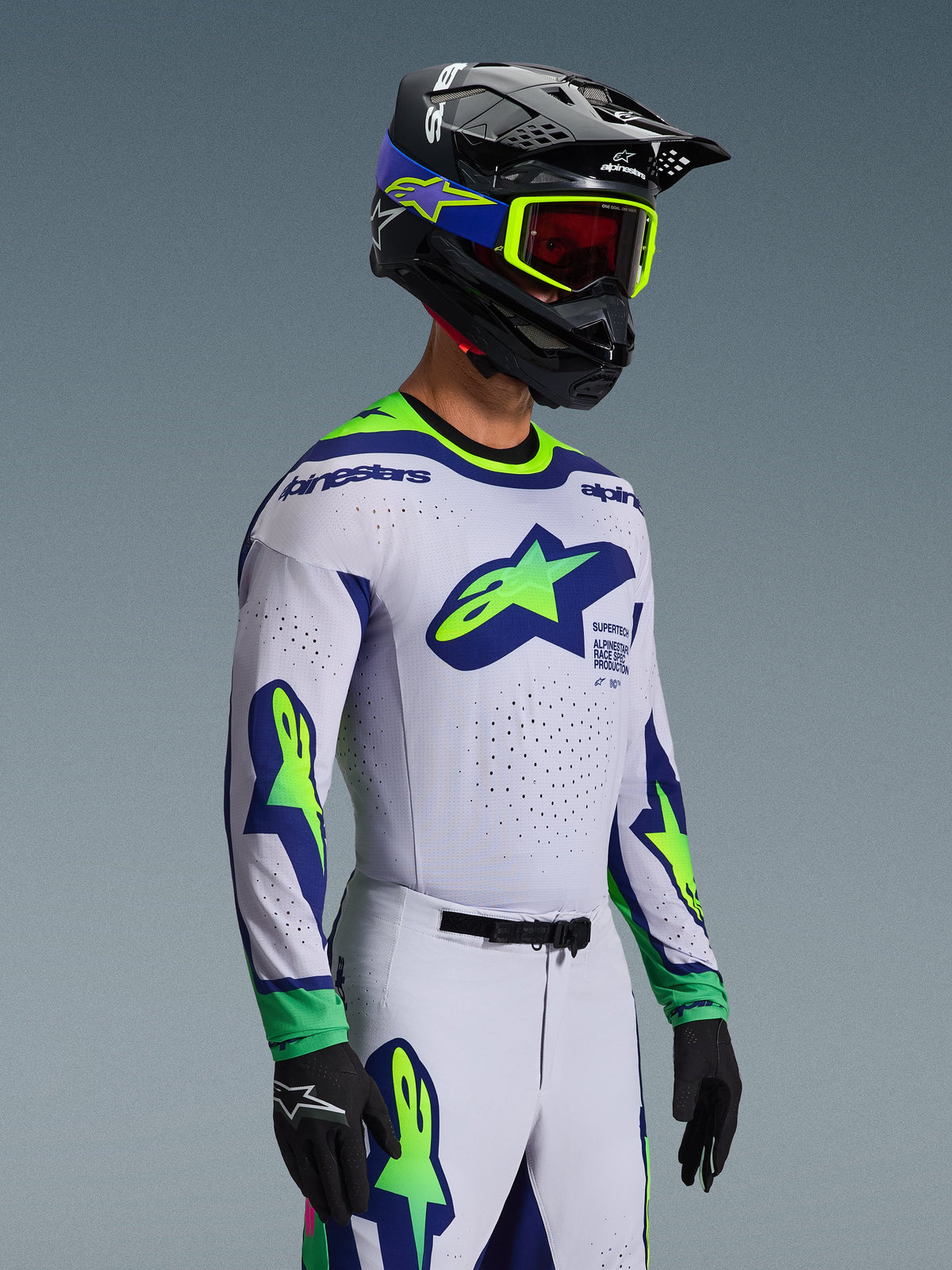 Completo Motocross Alpinestars Supertech Vista 2026, maglia e pantaloni, bianco con accenti blu e verde lime, indossato da un pilota con casco nero e occhiali verde neon, con loghi a stella prominenti sul petto e sulle maniche, vestibilità racing atletica di livello professionale per prestazioni off-road.