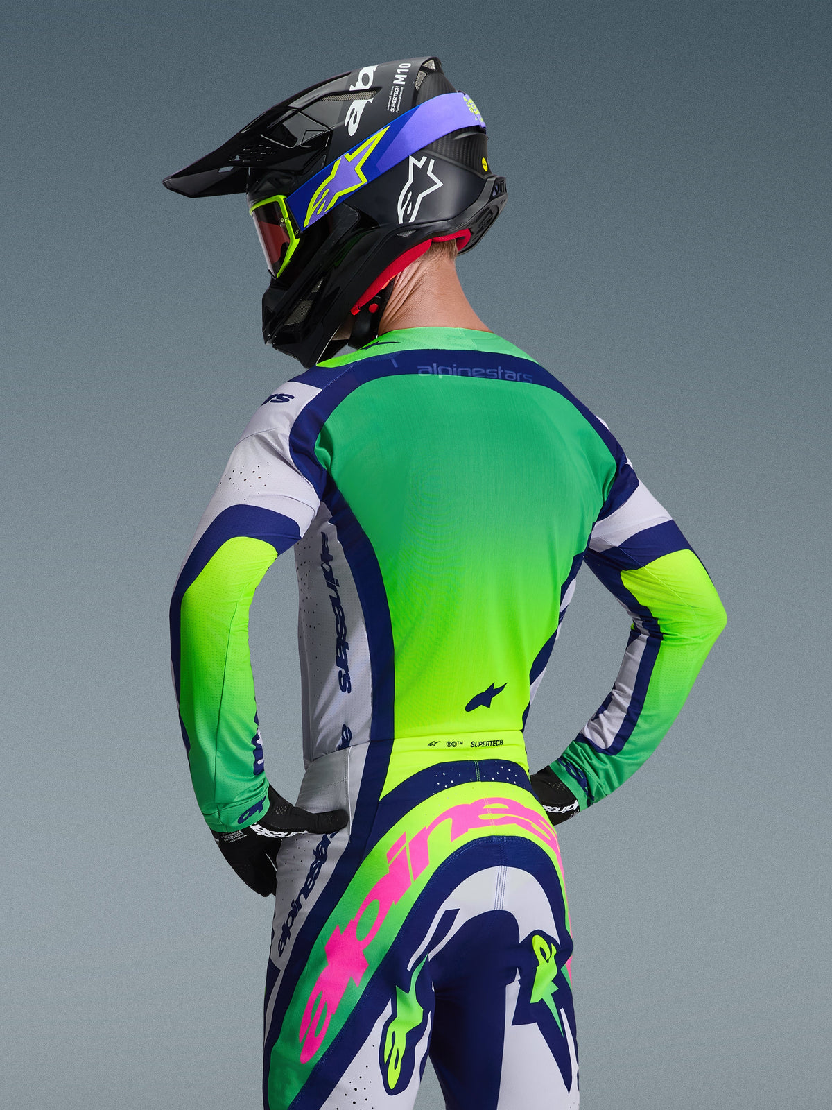 Vista posteriore di un pilota di motocross che indossa il completo Alpinestars 2026 Supertech Vista Gear Set, caratterizzato da una maglia verde e bianca con accenti blu navy, pantaloni coordinati con un prominente logo Alpinestars rosa, casco da corsa nero con grafiche blu e maschera con lenti rosse su uno sfondo grigio neutro.