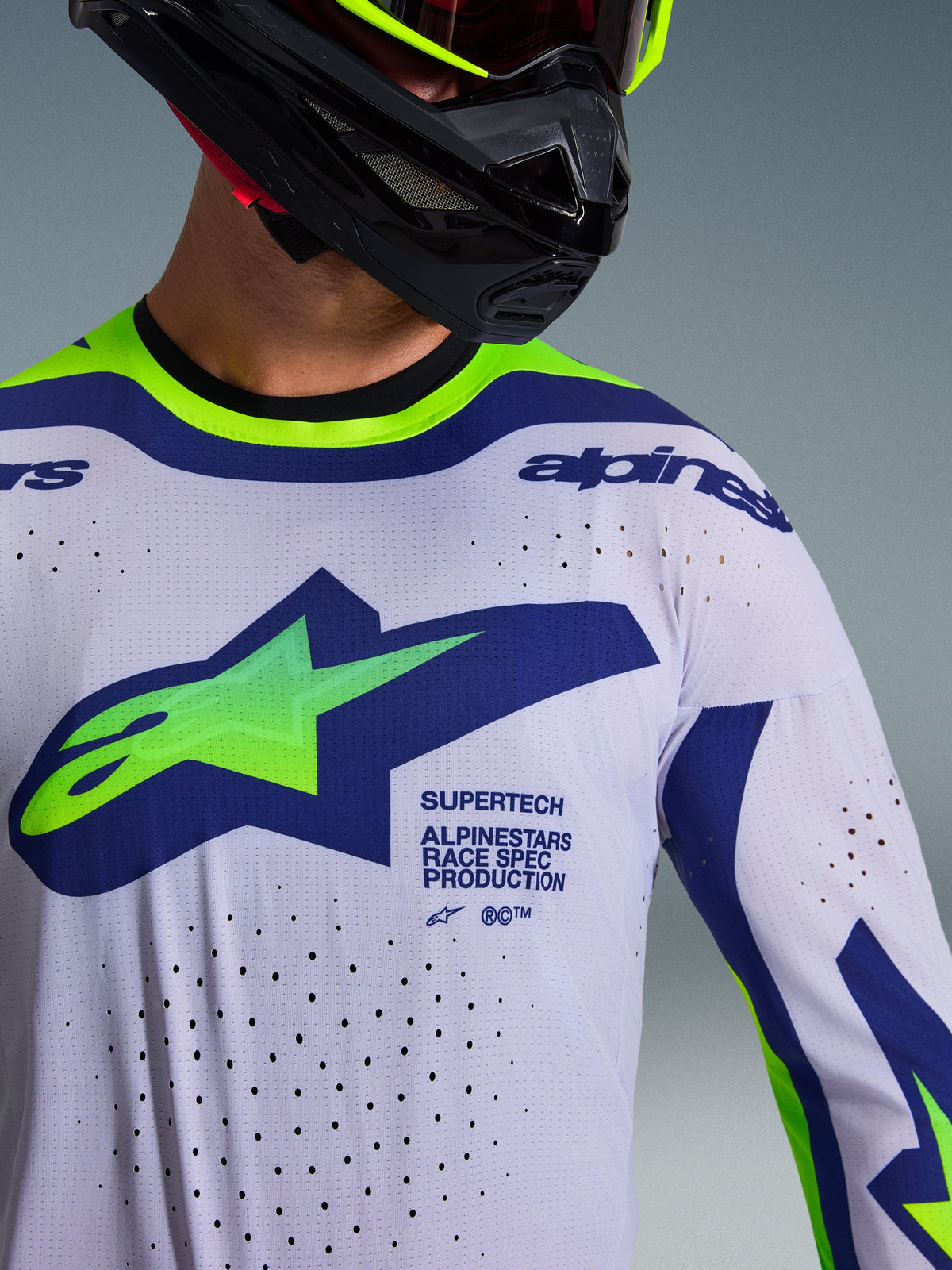 Maglia Alpinestars Supertech Vista 2026 in grigio chiaro, giallo fluo e blu, primo piano con grande logo sul petto e perforazioni laser per la ventilazione, indossata da un pilota con casco nero e branding professionale race-spec