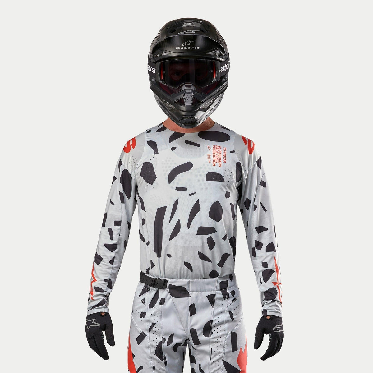 Maglia Alpinestars Techstar Rantera 2024, maglia da motocross, Haze Gray Camo con motivi neri irregolari e accenti rossi, indossata da un pilota con pantaloni coordinati, casco nero e guanti, equipaggiamento da gara fuoristrada.