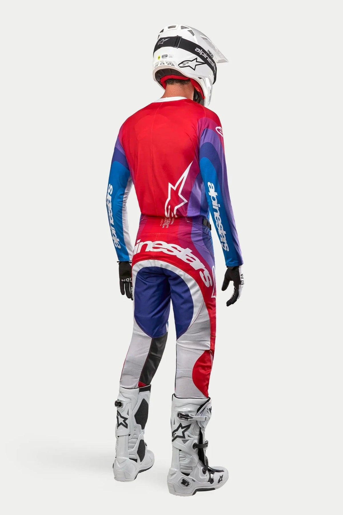 Pilota di motocross visto da dietro, maglia e pantaloni Alpinestars Techstar Pneuma 2024, colorazione Blu Mars Red White con sfumature rosse, blu e viola, loghi bianchi, stivali e casco bianchi, abbigliamento coordinato da gara fuoristrada