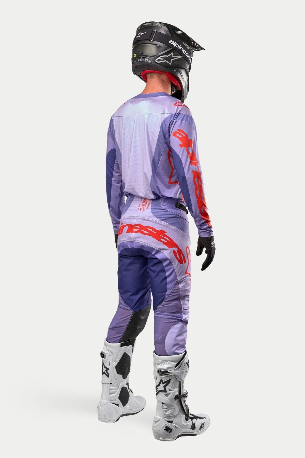 Pilota di motocross con abbigliamento da gara completo visto dal retro, maglia Alpinestars 2024 Techstar Ocuri viola con logo arancione acceso, pantaloni viola coordinati con dettagli arancioni, casco nero, stivali bianchi e neri, guanti neri, kit professionale da corsa fuoristrada