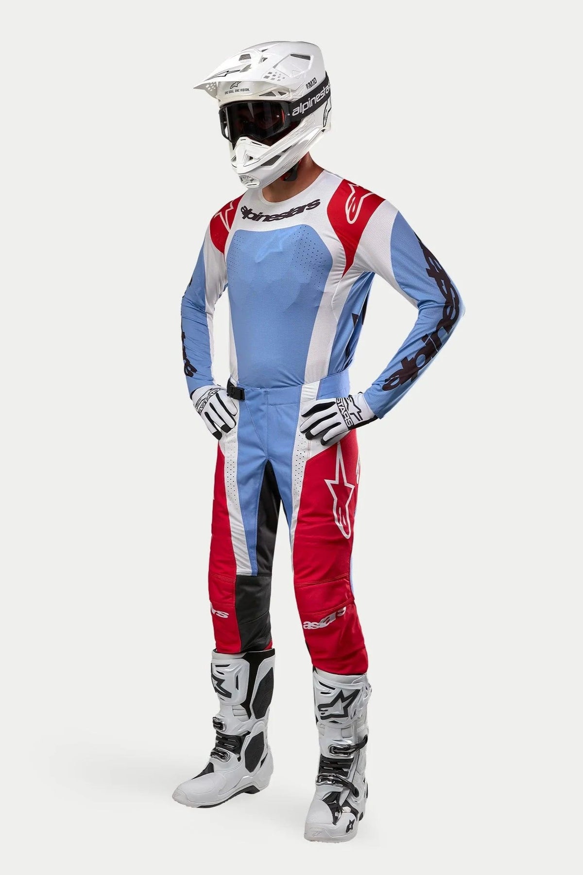 Pilota di motocross con equipaggiamento completo Alpinestars: maglia Techstar Ocuri 2024 in azzurro, rosso e bianco, pantaloni abbinati, casco bianco con occhiali, stivali bianchi e guanti bianchi e neri.