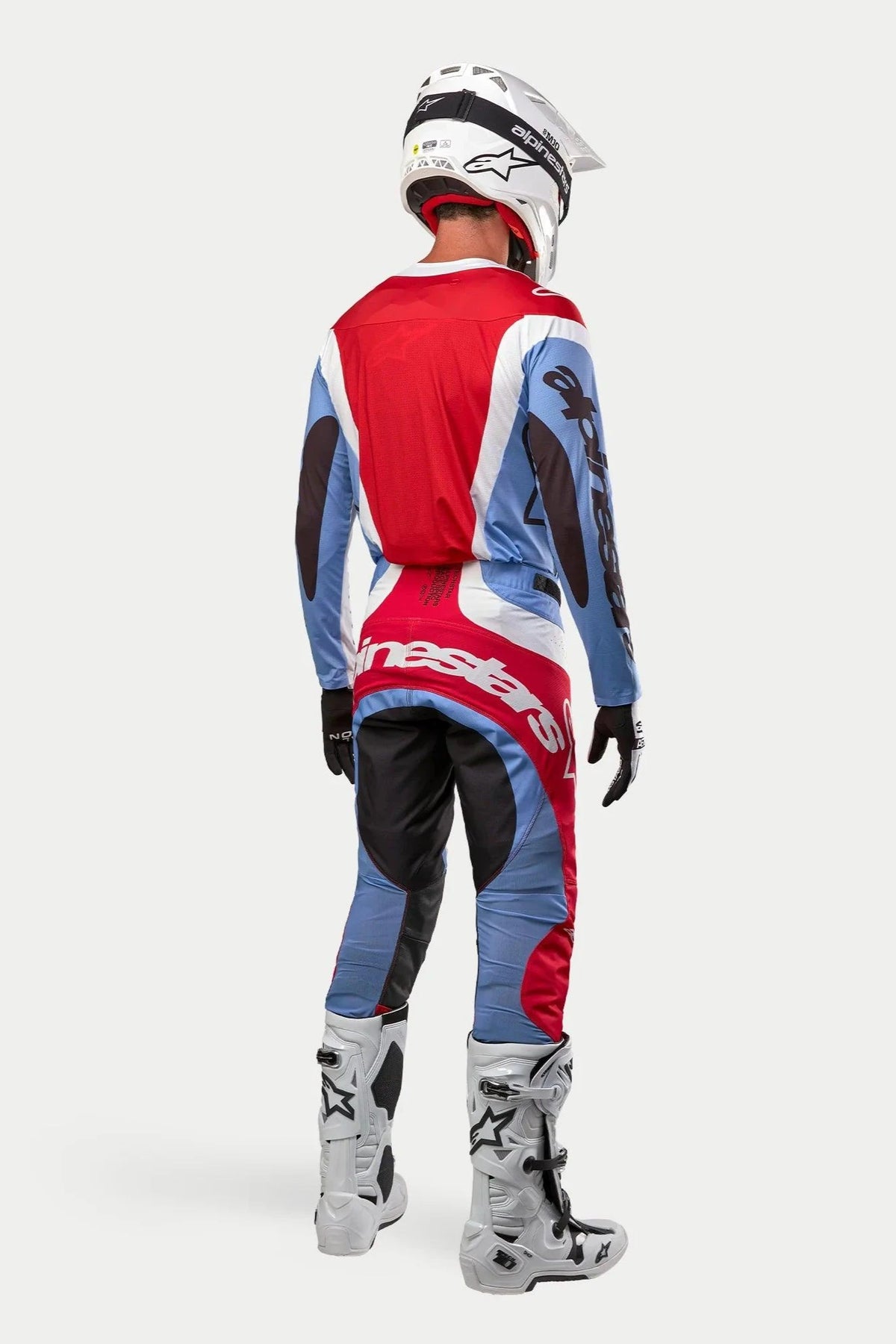 Pilota di motocross visto di spalle con equipaggiamento da gara completo, Maglia Techstar Ocuri 2024 in azzurro, rosso Marte e bianco, pantaloni coordinati con loghi evidenti, stivali bianchi e neri, casco bianco, completo coordinato per corse fuoristrada