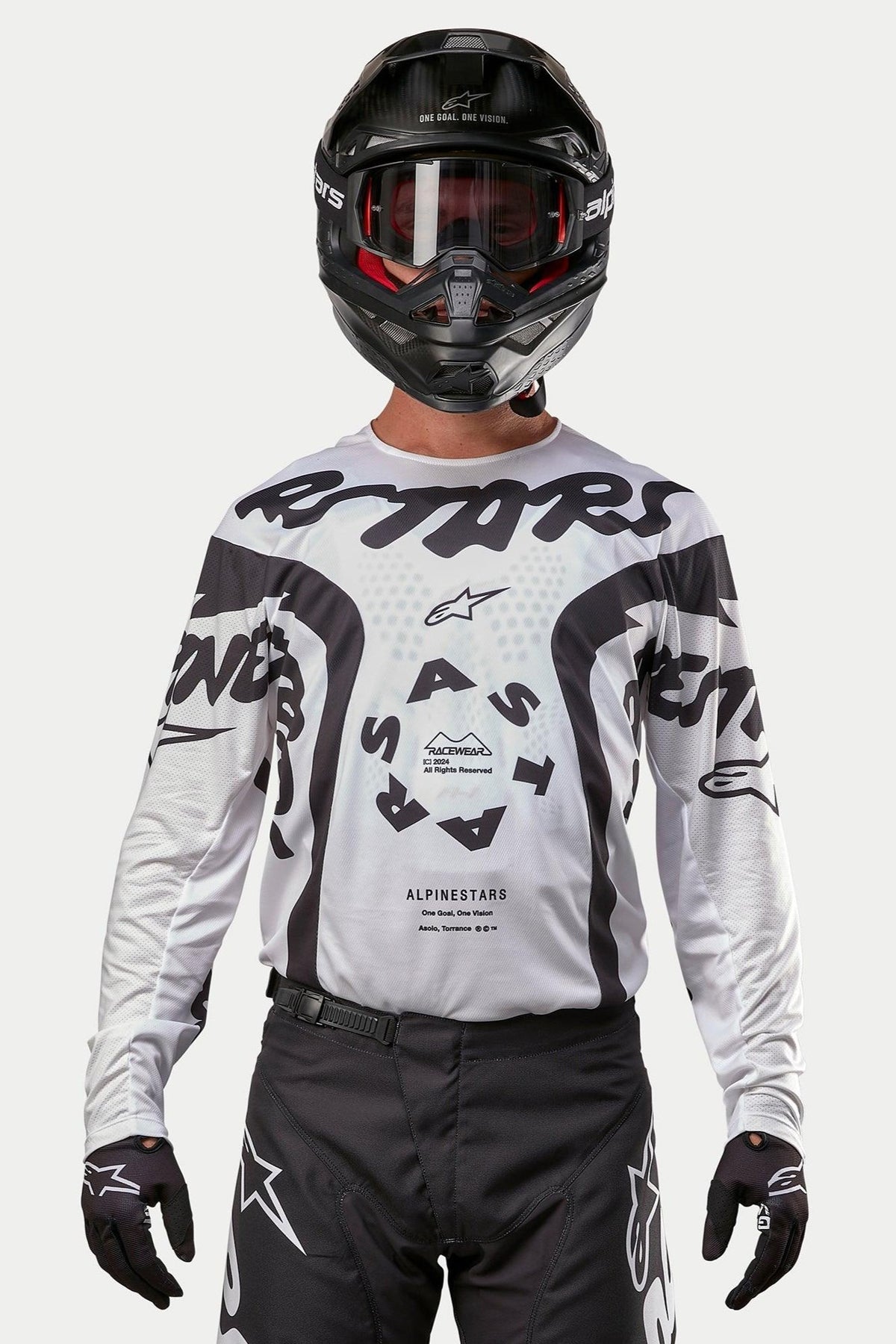 Pilota di motocross che indossa la maglia Alpinestars 2024 Racer Hana in bianco e nero, abbinata a un casco nero e pantaloni da motocross neri, con grafiche nere 'ASTARS' e un logo centrale circolare, equipaggiamento professionale per gare fuoristrada