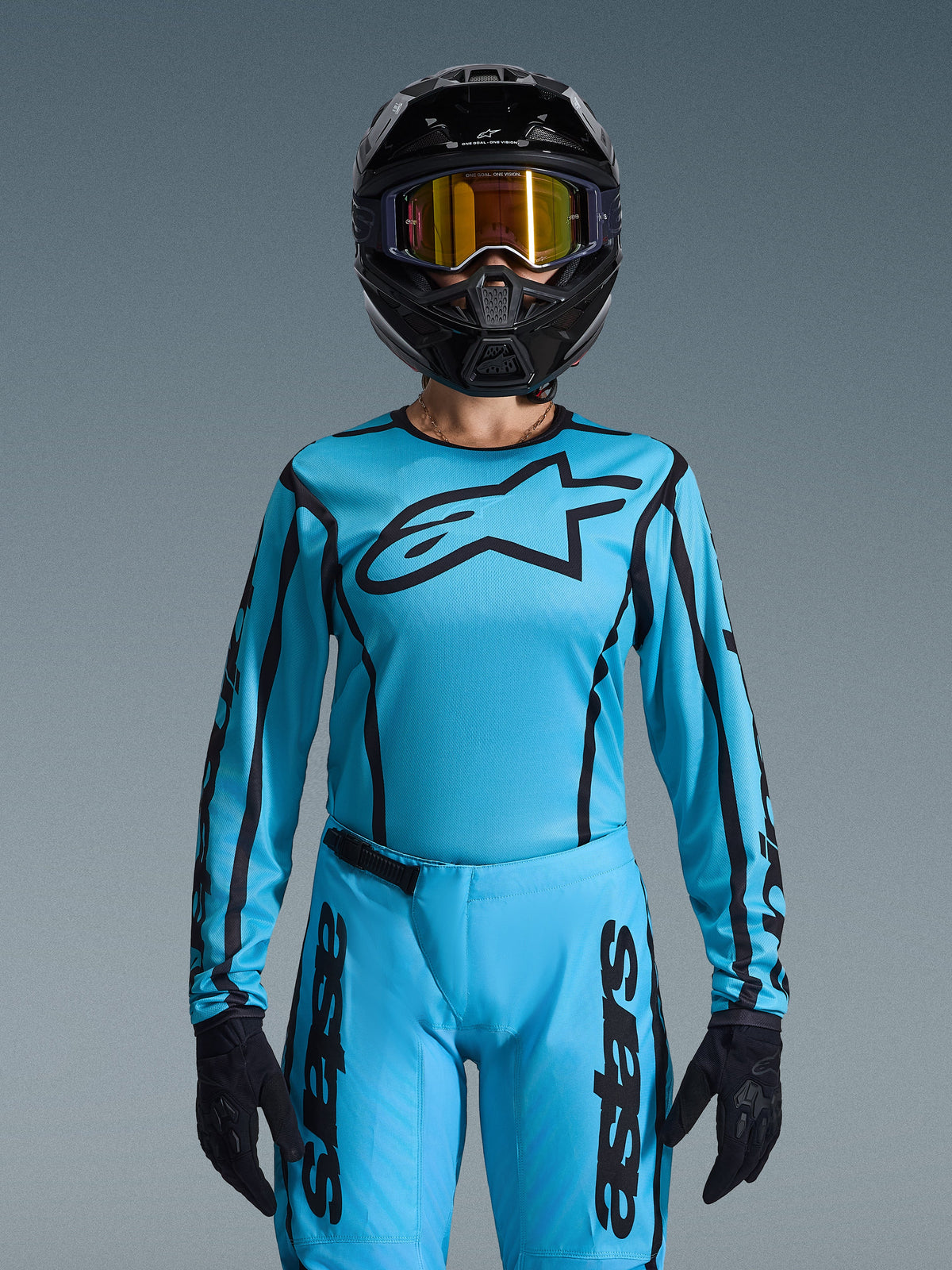 Pilota di motocross con completo Alpinestars Stella Fluid Apex 2026 da donna, maglia azzurra con contorni e logo neri, pantaloni coordinati, casco nero con maschera oro iridescente e guanti neri, abbigliamento da motocross femminile coordinato
