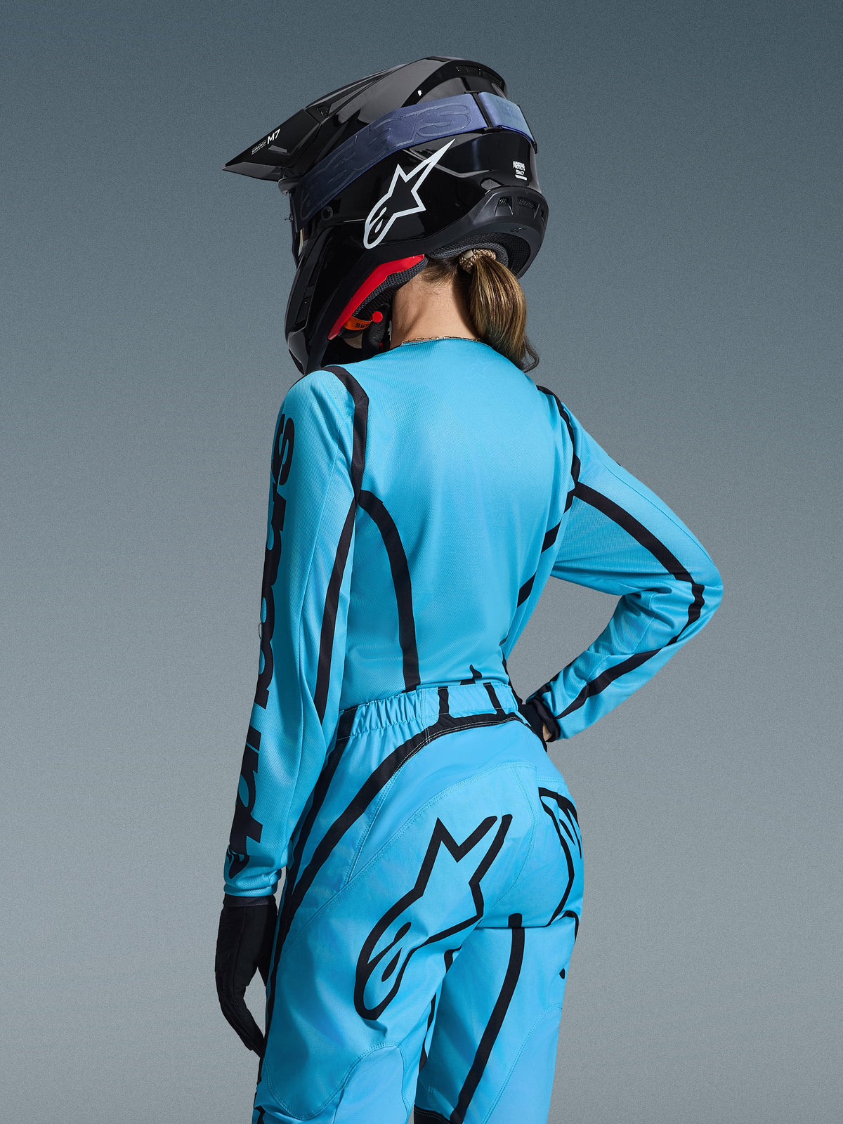 Completo Stella Fluid Apex Donna 2026 Alpinestars, azzurro con grafiche curve nere, vista posteriore su una pilota, indossato con casco e guanti neri, abbigliamento ergonomico da motocross per donna
