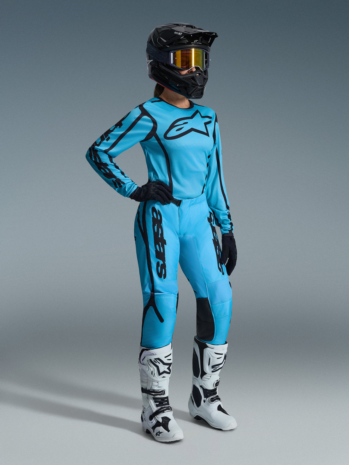Completo Alpinestars Stella Fluid Apex Donna 2026 in Tropical Black, maglia azzurra e pantaloni coordinati con dettagli neri, indossato da una pilota con casco nero, occhiali iridescenti e stivali bianchi, abbigliamento professionale per motocross