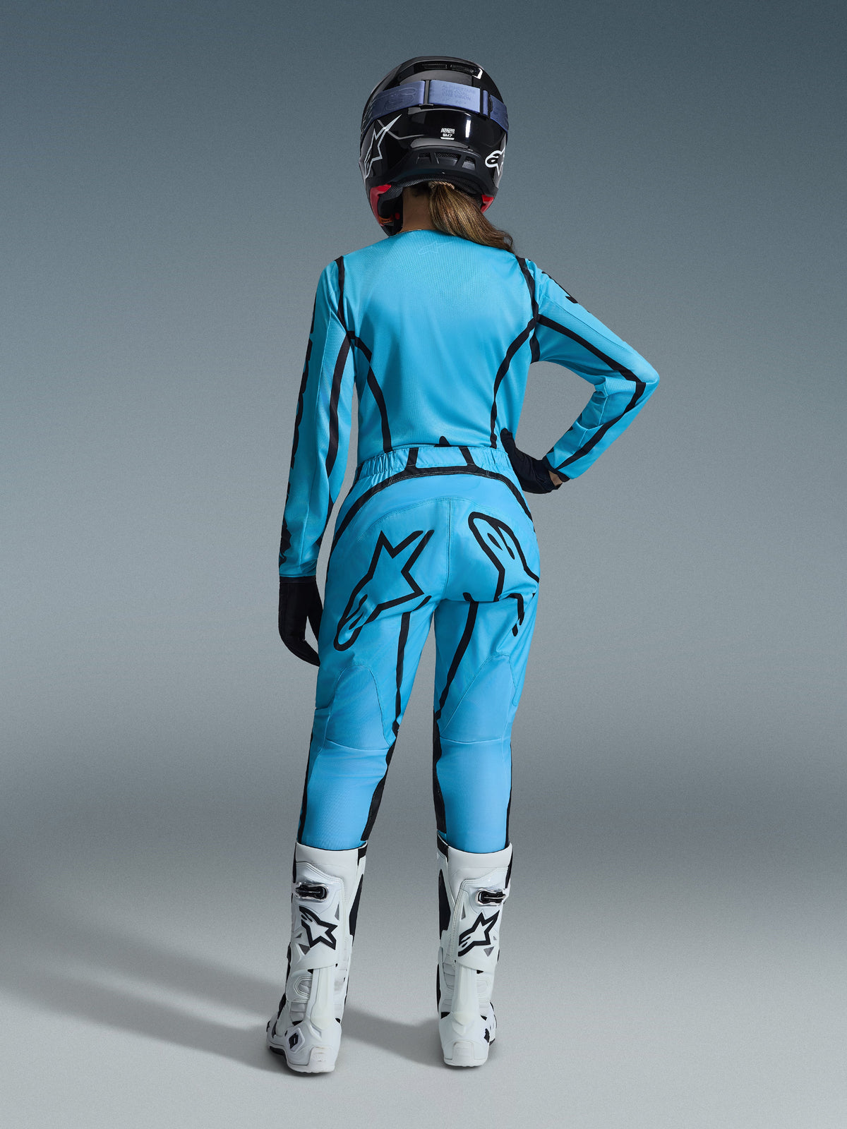 Pilota di motocross donna vista da dietro, che indossa il completo Alpinestars 2026 Woman Stella Fluid Apex Gear Set in blu acceso con accenti grafici a linee nere, caratterizzato da un grande logo a stella nero sui pantaloni, abbinato a stivali bianchi e neri e un casco nero.