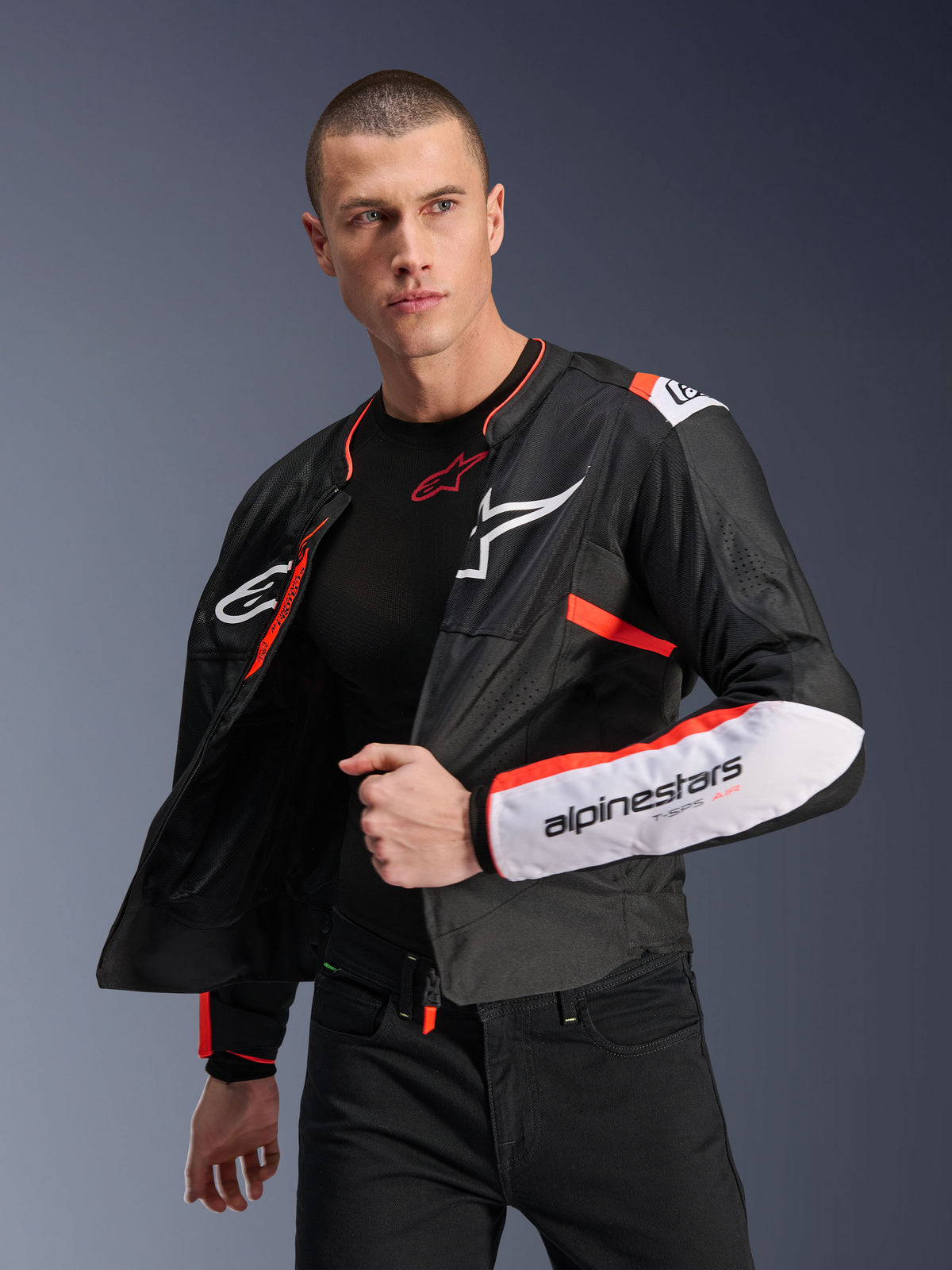 Modello che indossa la Giacca Alpinestars T-SPS Air V2, giacca da moto in tessuto nero con accenti bianchi e rosso fluorescente, pannelli in mesh per il flusso d'aria, logo Alpinestars bianco sul petto e marchio sulla manica, indossata aperta sopra un base layer nero