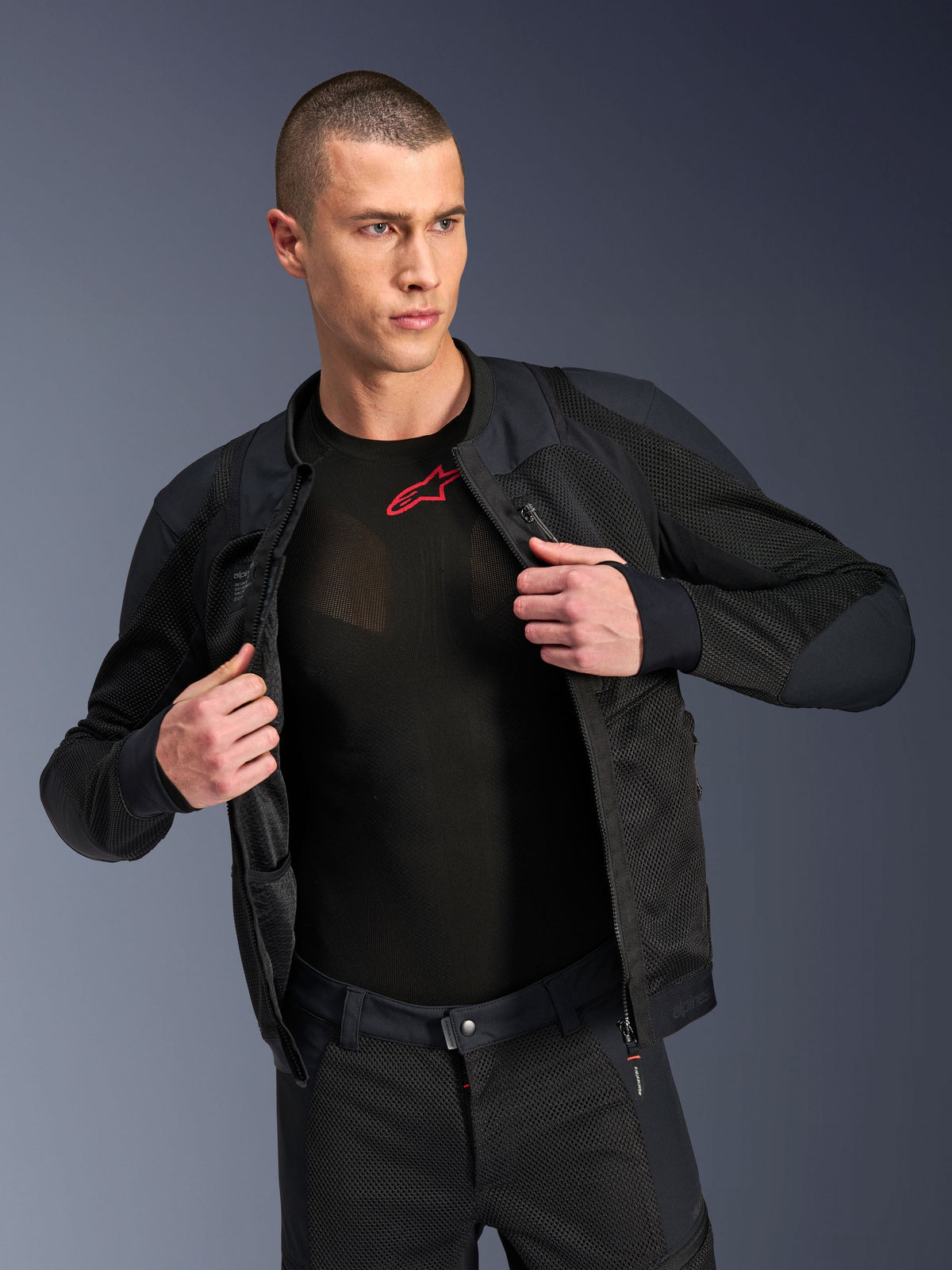 Giacca Alpinestars Troop-Air Pro, nera, giacca tecnica protettiva ventilata di alta qualità con struttura in mesh, indossata aperta su un base layer nero con logo rosso, abbinata a pantaloni da moto in mesh coordinati, stile di guida urban contemporary