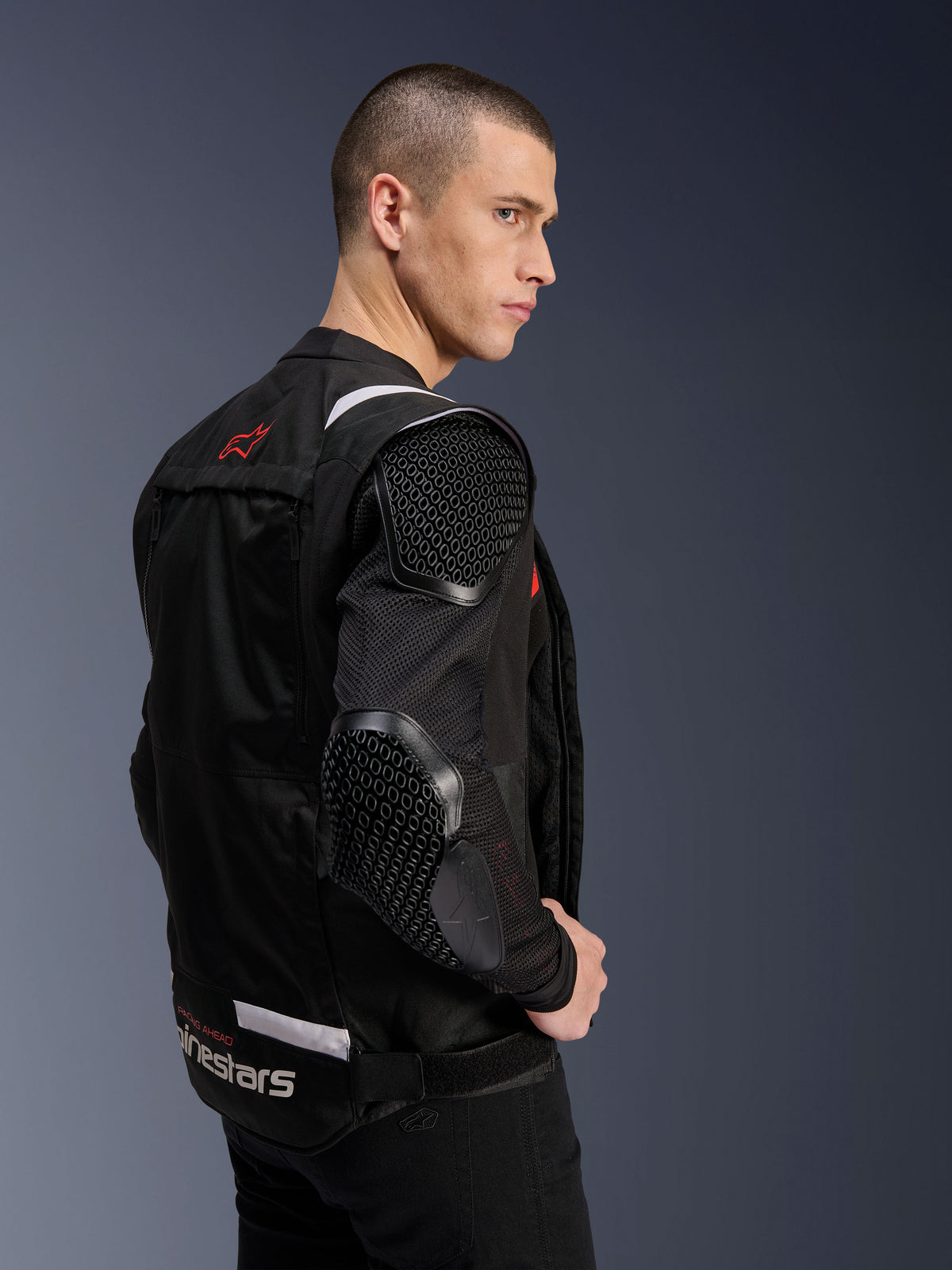 Giacca Alpinestars T-Stunt Air, giacca da moto, nero, profilo laterale di un modello che indossa la giacca dal taglio sportivo con maniche in rete e protezioni con motivo a nido d'ape visibili su spalla e gomito, logo rosso sulla schiena, branding bianco in vita