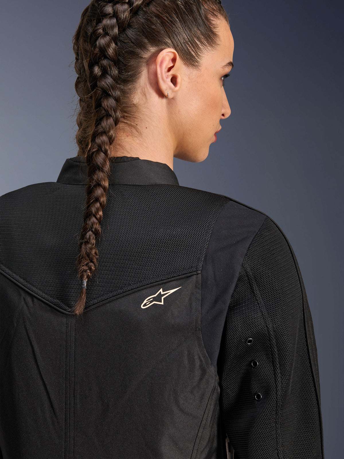 Alpinestars Stella T-Dyno Air Jacket, giacca da moto da donna nera, vista posteriore ravvicinata con pannelli in rete 3D sullo sprone e sulla parte superiore della schiena per la ventilazione, piccolo logo a stella bianco, indossata da una modella con capelli intrecciati.
