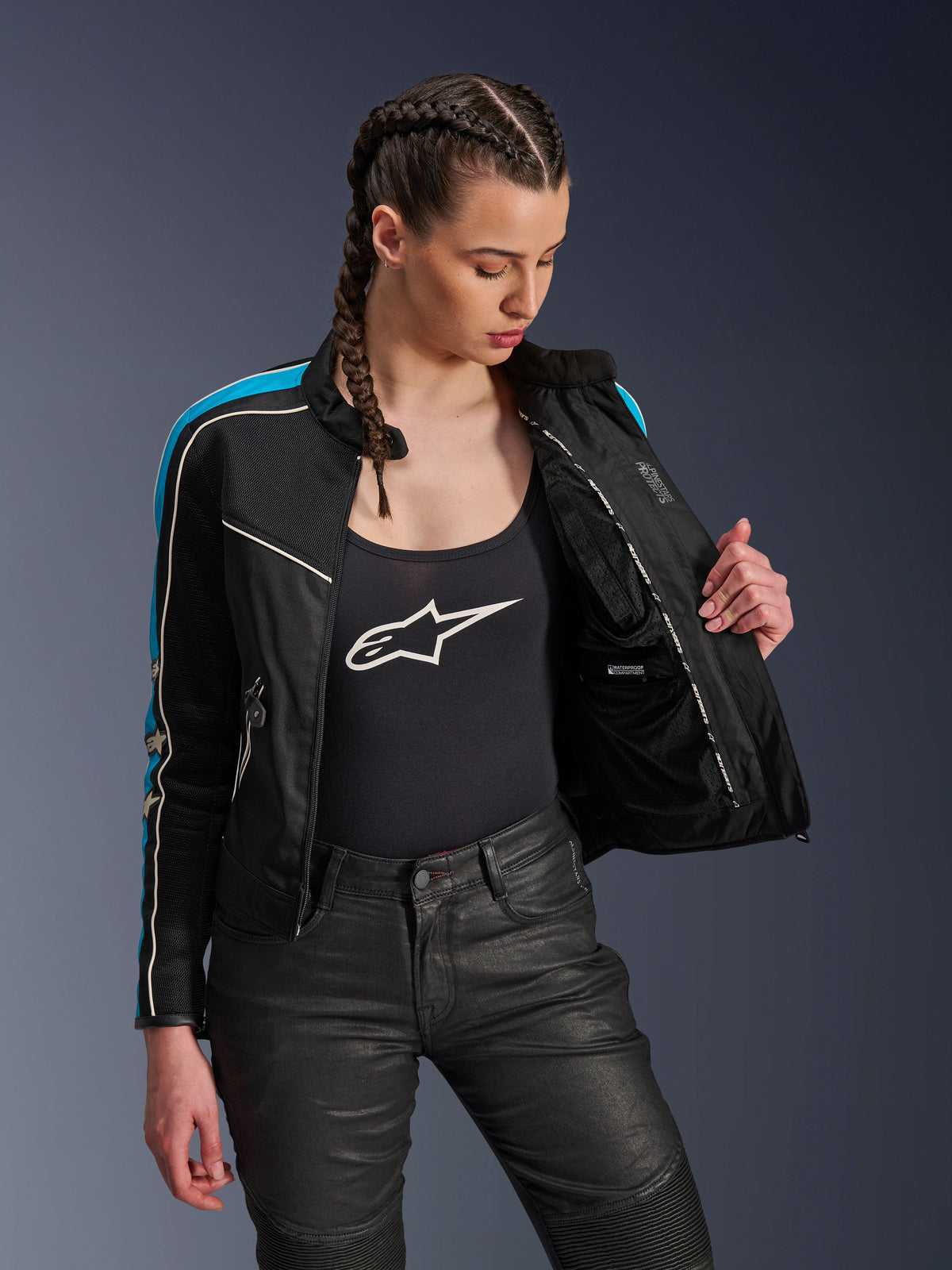 Donna che indossa la giacca Alpinestars Stella T-Dyno Air in Black Light Blue Ecru, giacca aperta per mostrare la fodera interna in rete e la tasca impermeabile, abbinata a canotta nera con logo e pantaloni da moto neri, abbigliamento urbano d'ispirazione vintage