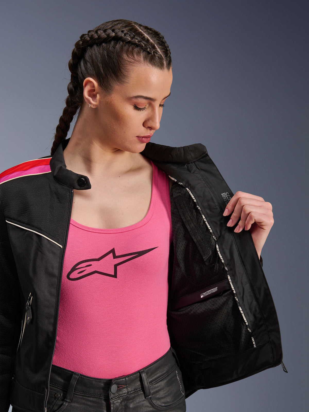 Motociclista che indossa la giacca Alpinestars Stella T-Dyno Air in nero, rosso acceso e fucsia, mostrata parzialmente aperta per rivelare lo scomparto interno impermeabile, indossata sopra una canotta rosa con logo, mettendo in risalto i pannelli in rete traspirante e la vestibilità sartoriale da motociclista.
