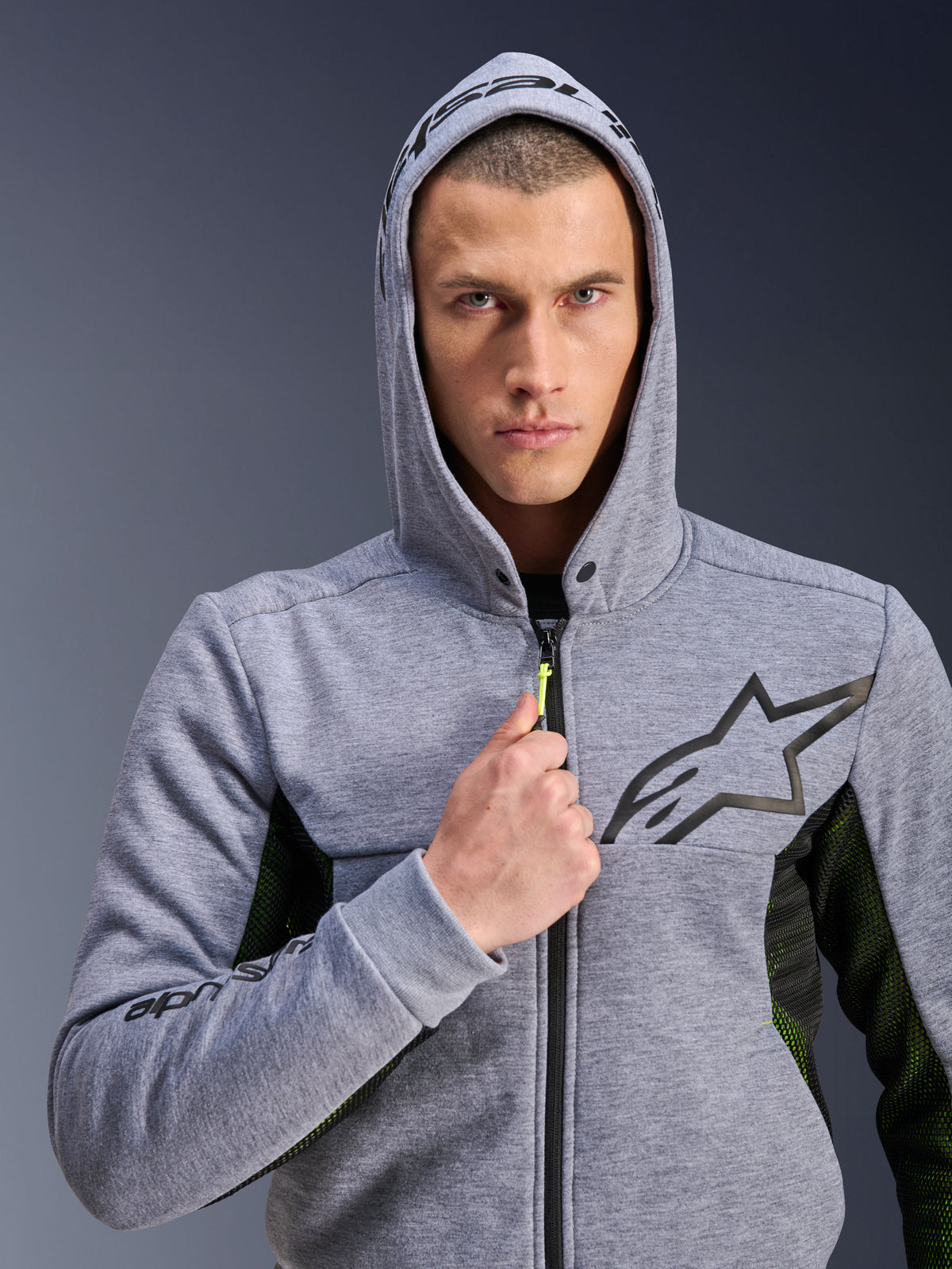 Alpinestars Chrome Air Sport Hoodie, Grigio Melange Nero, felpa da moto indossata da un uomo con il cappuccio alzato, caratterizzata da un logo a stella nero sul petto, tirante della zip giallo neon e pannelli laterali in mesh per la ventilazione, stile urban riding