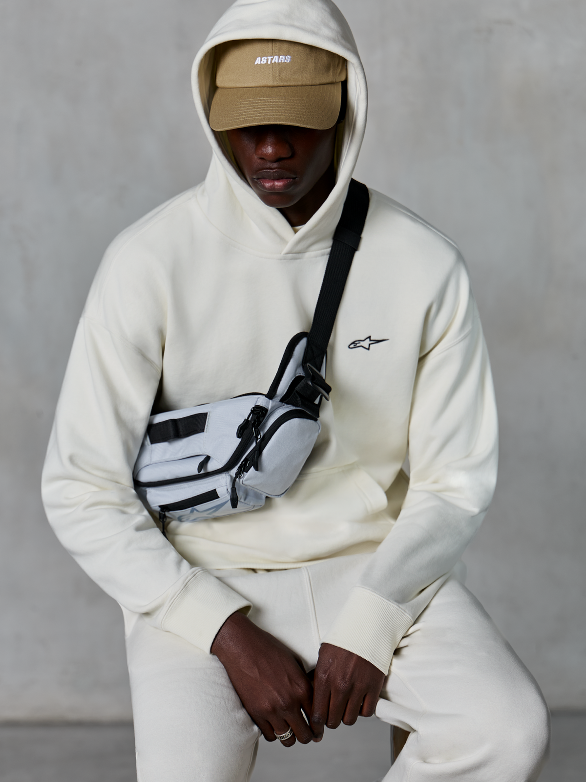 Alpinestars Essentials Ageless Hoodie, streetwear casual, Off White, piccolo logo nero ricamato sul petto, cappuccio indossato sopra un berretto color sabbia con marchio ASTARS, abbinata a una borsa a tracolla grigio chiaro, abbigliamento lifestyle premium