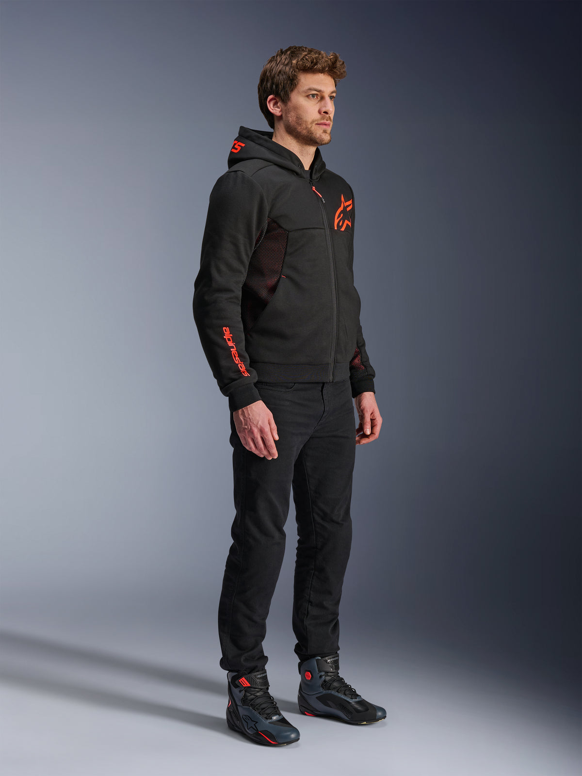 Alpinestars Chrome Air Sport Hoodie, Nero Rosso Fluo, giacca da guida urbana in pile tecnico, indossata con pantaloni neri e stivali da moto, con pannelli di ventilazione in mesh e loghi rossi fluorescenti sul petto e sulla manica.