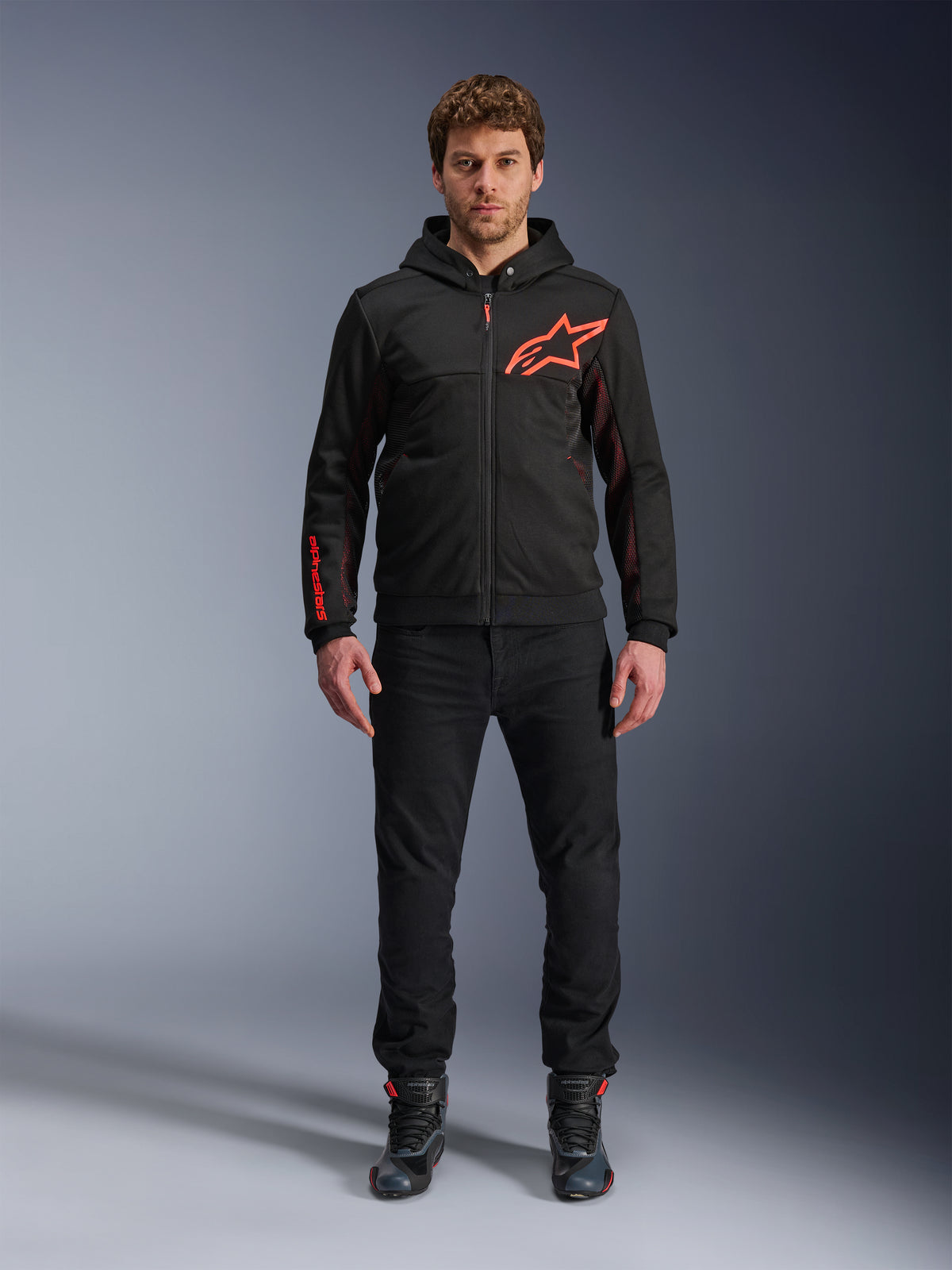 Alpinestars Chrome Air Sport Hoodie, nero e rosso fluo, giacca da moto urbana in pile tecnico, con grande logo Astars rosso sul petto e marchio sulla manica, indossata da un modello con pantaloni neri e scarpe da moto