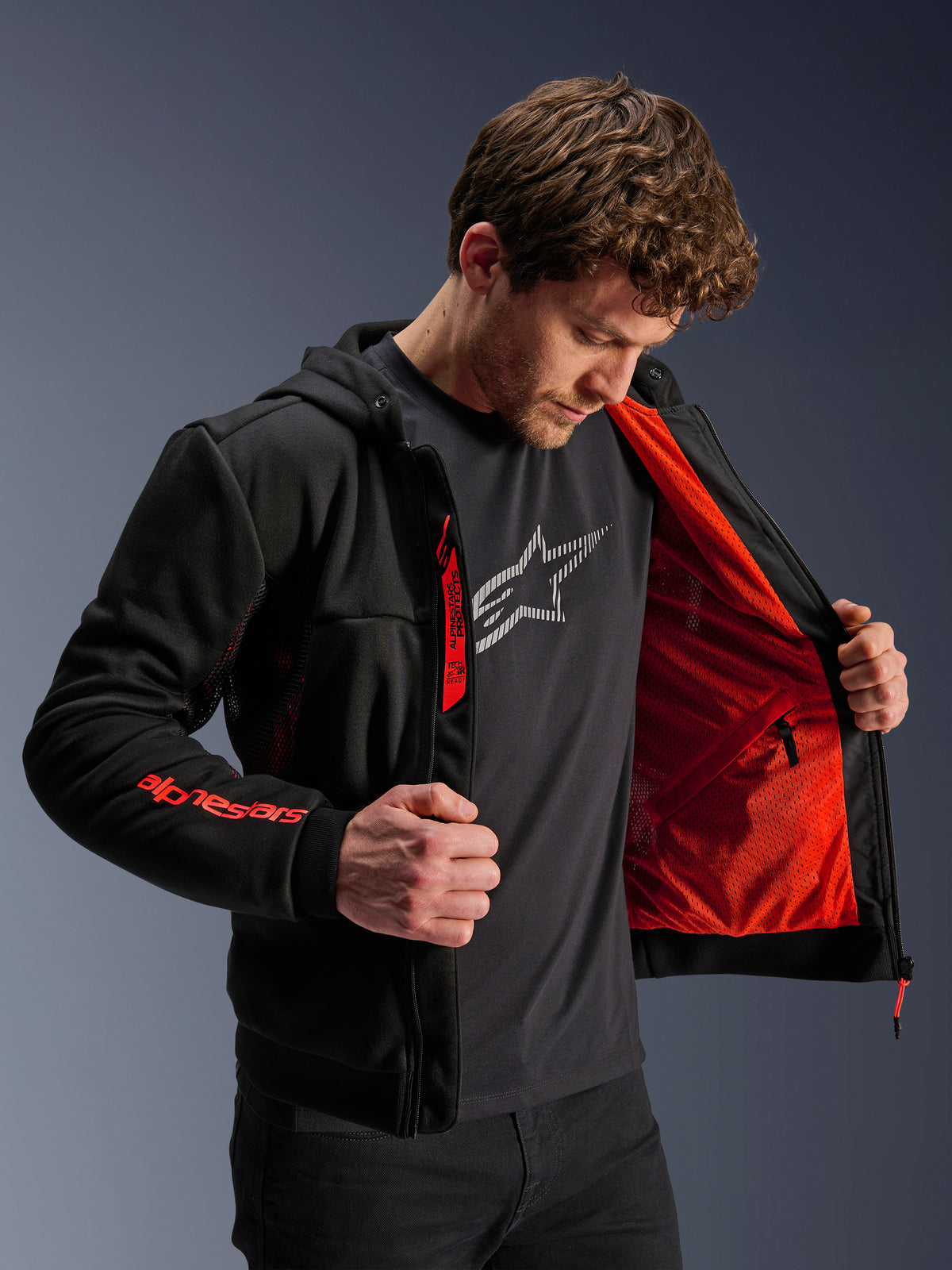 Alpinestars Chrome Air Sport Hoodie in nero e rosso fluo, modello maschile che apre la giacca per mostrare la fodera in mesh rosso brillante, branding Alpinestars sulla manica destra e sulla cerniera del petto, giacca da moto urbana casual progettata per il flusso d'aria.