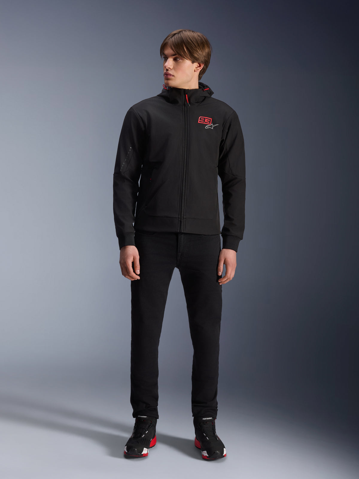 Alpinestars MM93 Radium Tech Hoodie, felpa da moto urbana nera, con logo 93 rosso e logo a stella bianco sul petto, indossata da un modello con pantaloni neri e scarpe da guida coordinate, guscio tecnico antivento per la guida urbana