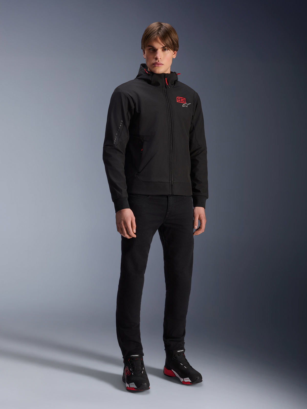 Alpinestars MM93 Radium Tech Hoodie, giacca da moto urban, nero con accenti rosso brillante, indossata da un modello con pantaloni neri e scarpe da moto, con logo '93' rosso sul petto e marchio Marc Marquez sulla manica, streetwear tecnico casual per la guida urbana