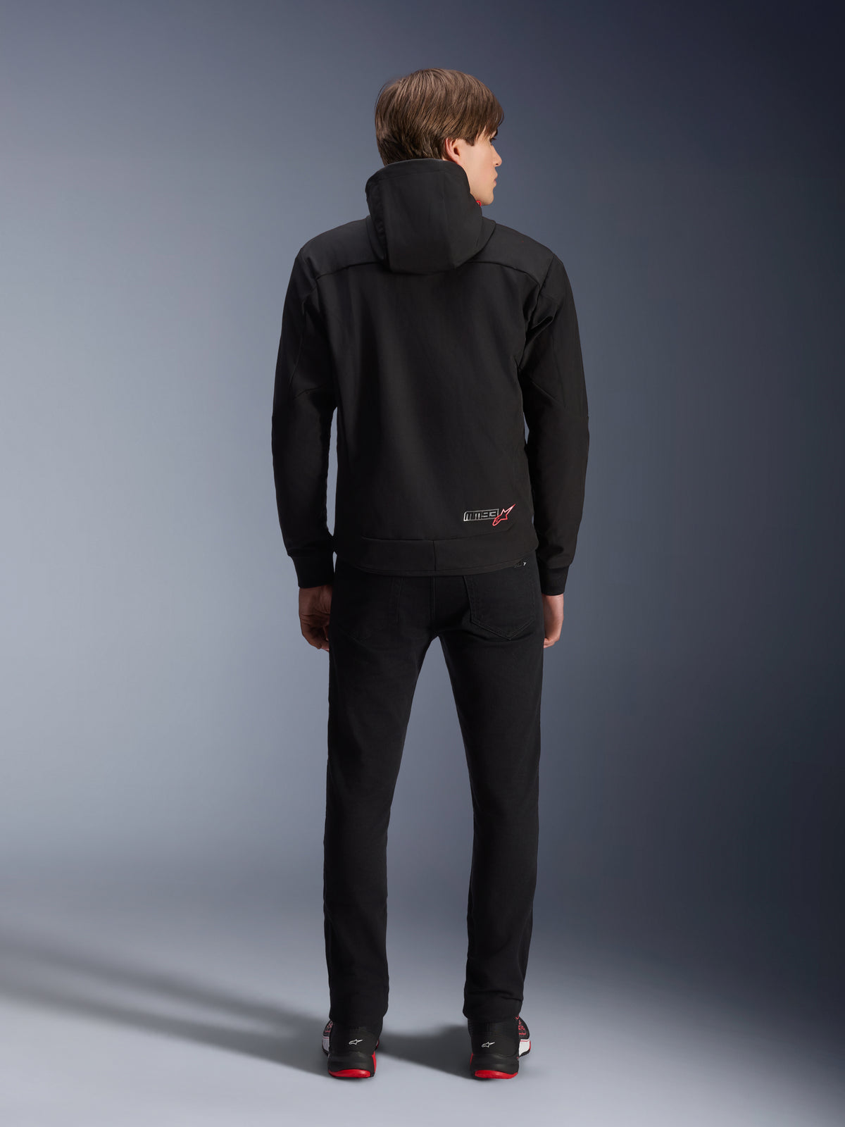 Alpinestars MM93 Radium Tech Hoodie, nera, vista posteriore su modello, con un piccolo logo MM93 e stella rossa sulla parte inferiore della schiena, abbinata a pantaloni neri, abbigliamento per la guida urbana in moto