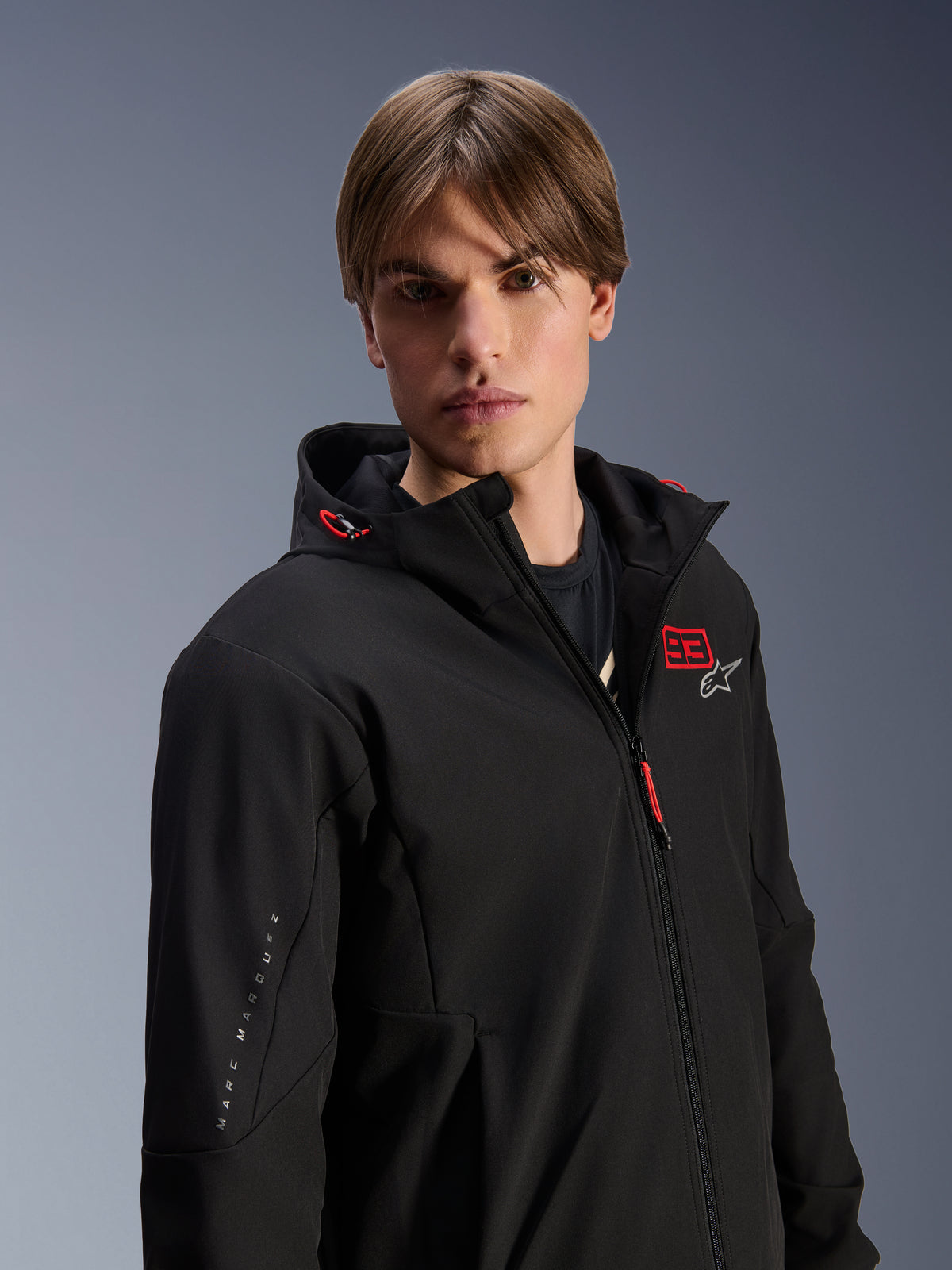 Modello che indossa la felpa Alpinestars MM93 Radium Tech Hoodie, giacca da moto urbana nera con logo '93' rosso e stella bianca sul petto, marchio 'Marc Marquez' sulla manica, guscio tecnico antivento per la guida urbana