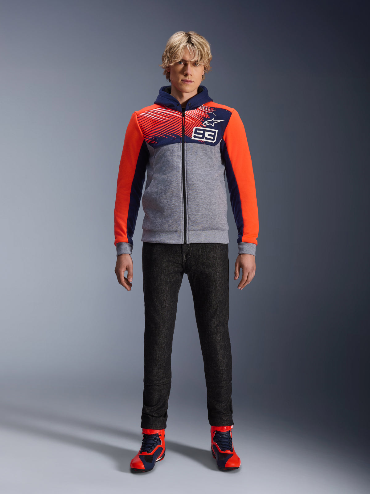 Alpinestars MM93 Chrome V2 Sport Hoodie, felpa da moto, blu navy, grigio melange e rosso fluo, indossata con pantaloni da guida scuri e sneaker con accenti rossi, dotata di cerniera integrale e logo MM93, design casual d'ispirazione motorsport urbana