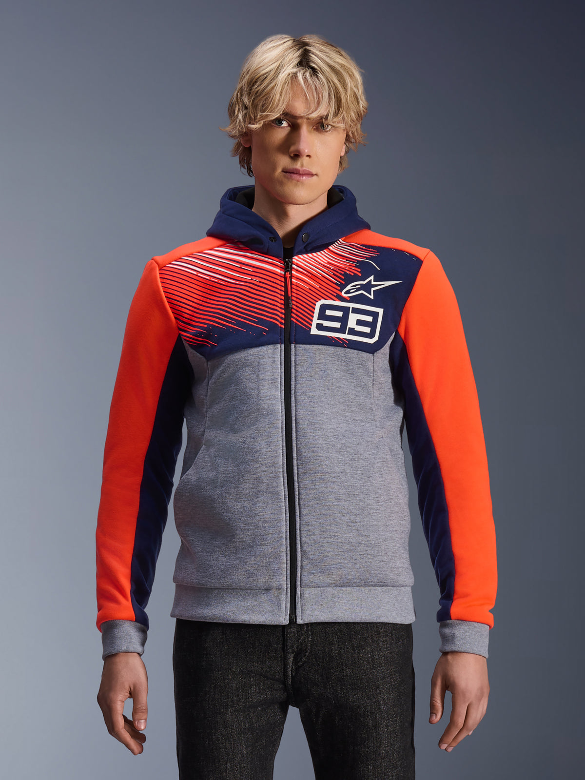 Alpinestars MM93 Chrome V2 Sport Hoodie, felpa da moto con zip, grigio mélange con petto e cappuccio blu navy, maniche arancione acceso, design con numero 93 sul petto con motivo a strisce e logo a stella, indossata da un modello con denim scuro, Collezione Marc Marquez.
