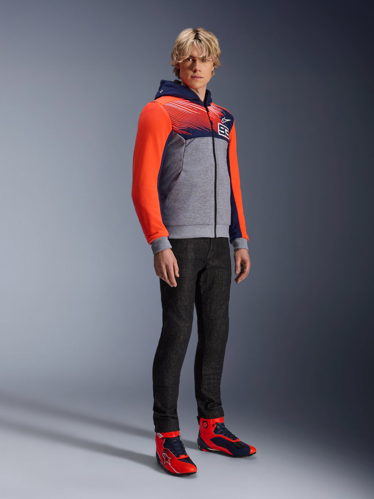 Alpinestars MM93 Chrome V2 Sport Hoodie, felpa da moto urbana, corpo grigio melange con petto blu navy e maniche rosso fluo acceso, con logo 93 sul petto, indossata da un modello con pantaloni neri slim-fit e scarpe da moto alte coordinate rosse e blu, vestibilità atletica moderna per la guida urbana