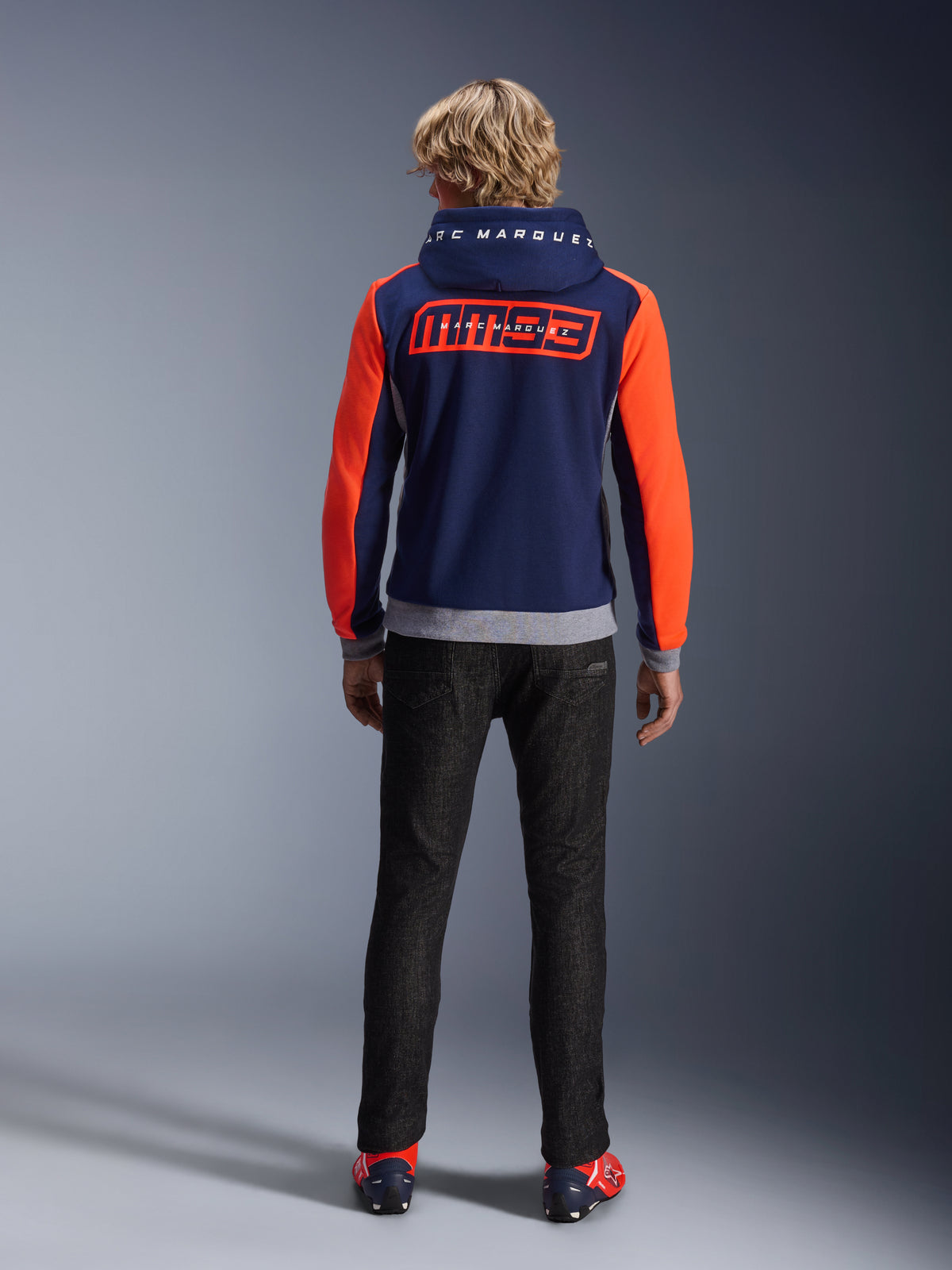 Modello visto di spalle che indossa la felpa Alpinestars MM93 Chrome V2 Sport Hoodie, blu navy con maniche rosso fluo e dettagli grigio melange, grandi loghi MM93 e Marc Marquez sulla schiena e sul cappuccio, felpa da moto casual abbinata a pantaloni neri