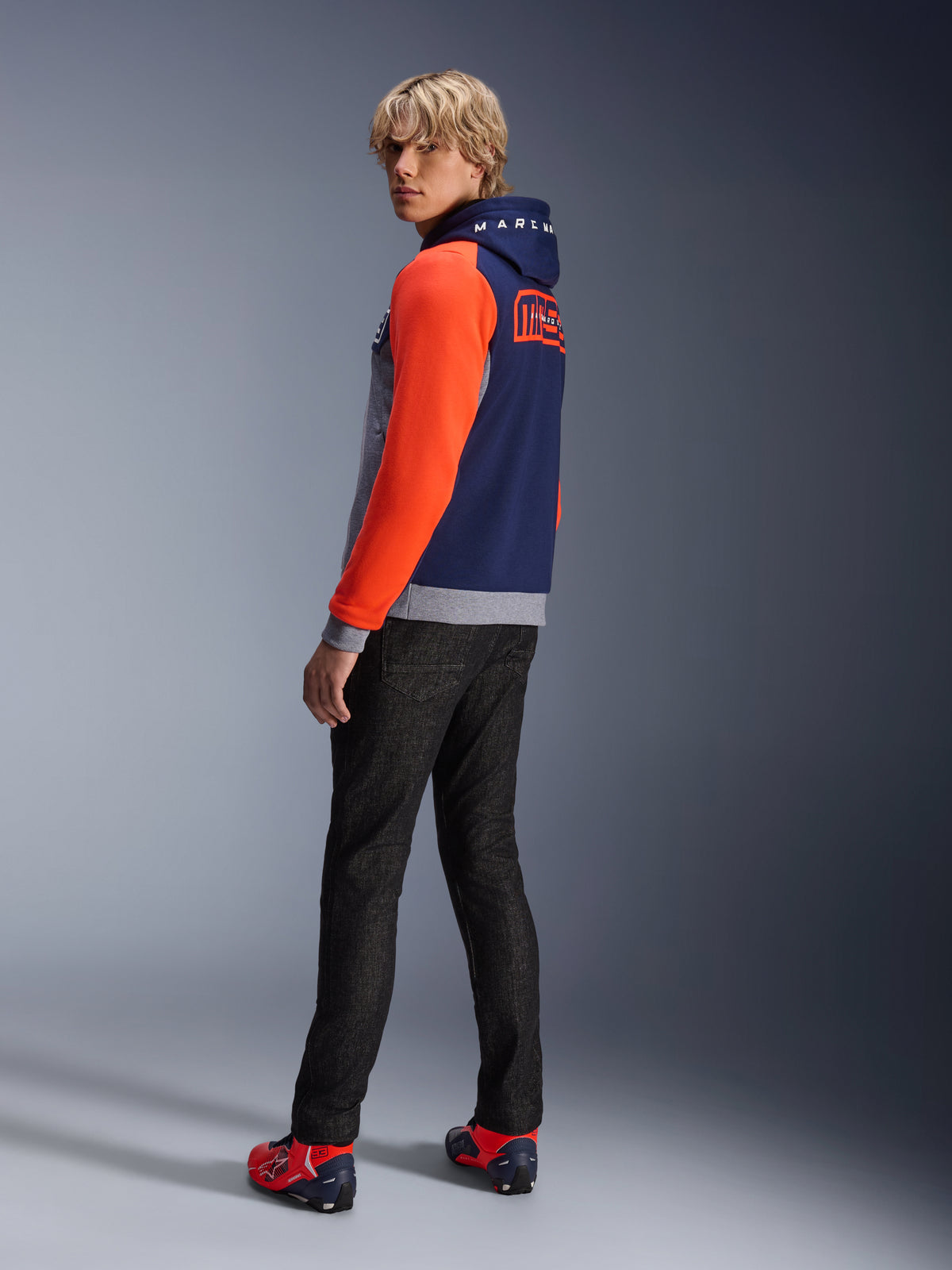Alpinestars MM93 Chrome V2 Sport Hoodie, Blue Navy Grigio Melange Rosso Fluoro, modello che indossa la felpa con maniche rosso acceso e branding MM93 Marc Marquez sul retro, abbinata a jeans scuri e scarpe da moto rosse, abbigliamento moto urban casual