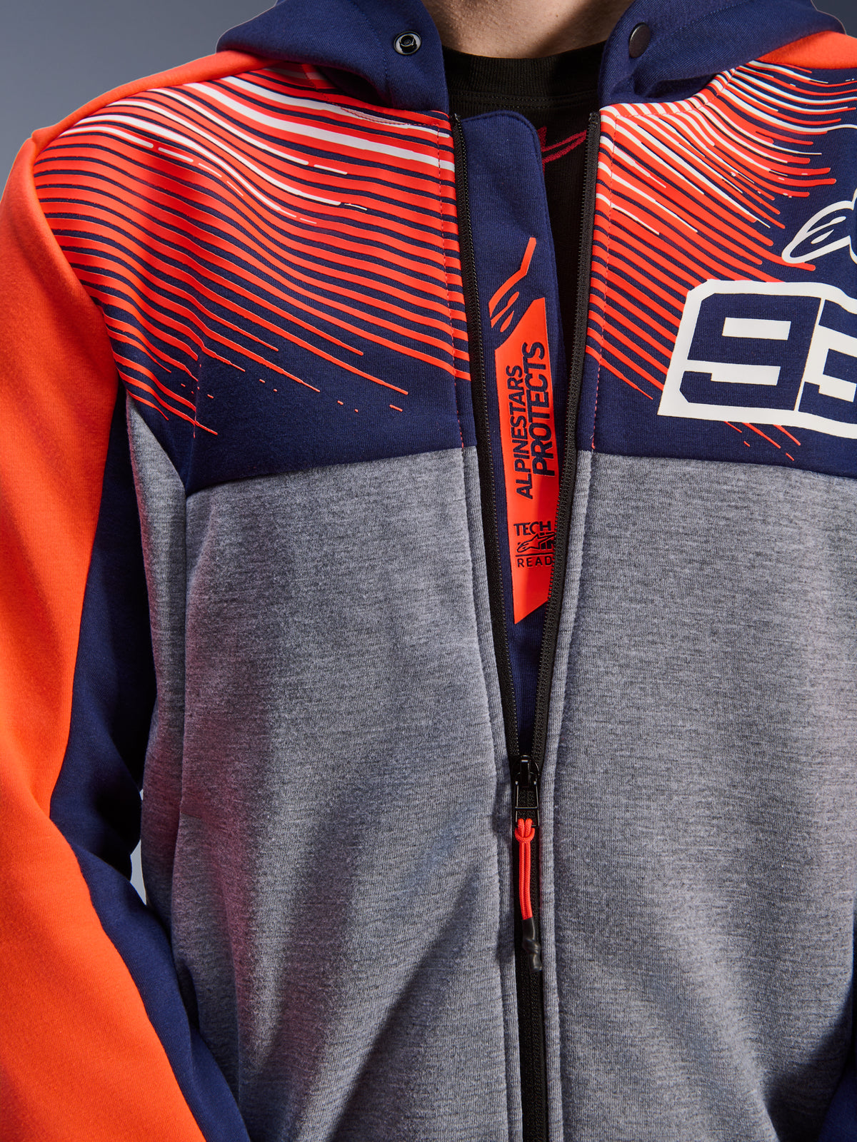 Alpinestars MM93 Chrome V2 Sport Hoodie, primo piano del petto e del colletto, pile tecnico blu navy e grigio melange con grafiche rosso fluo, logo 93 bianco e branding Alpinestars Protects sulla patta interna della cerniera, abbigliamento da moto Tech-Air Ready