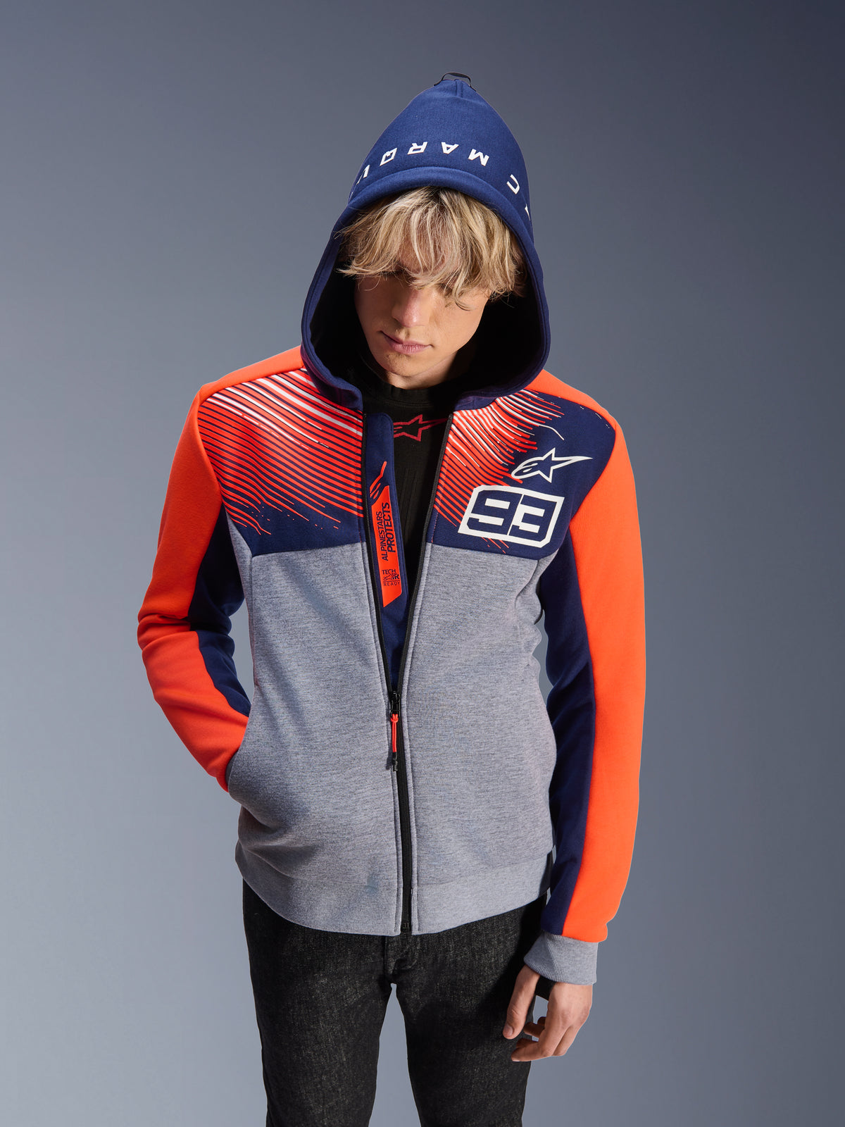 Alpinestars MM93 Chrome V2 Sport Hoodie, felpa da moto, corpo grigio melange con parte superiore blu navy e maniche arancione fluorescente, con numero 93 bianco e logo Alpinestars, scritta Marc Marquez sul cappuccio, indossata da un modello con cappuccio alzato, design d'ispirazione urban motorsport
