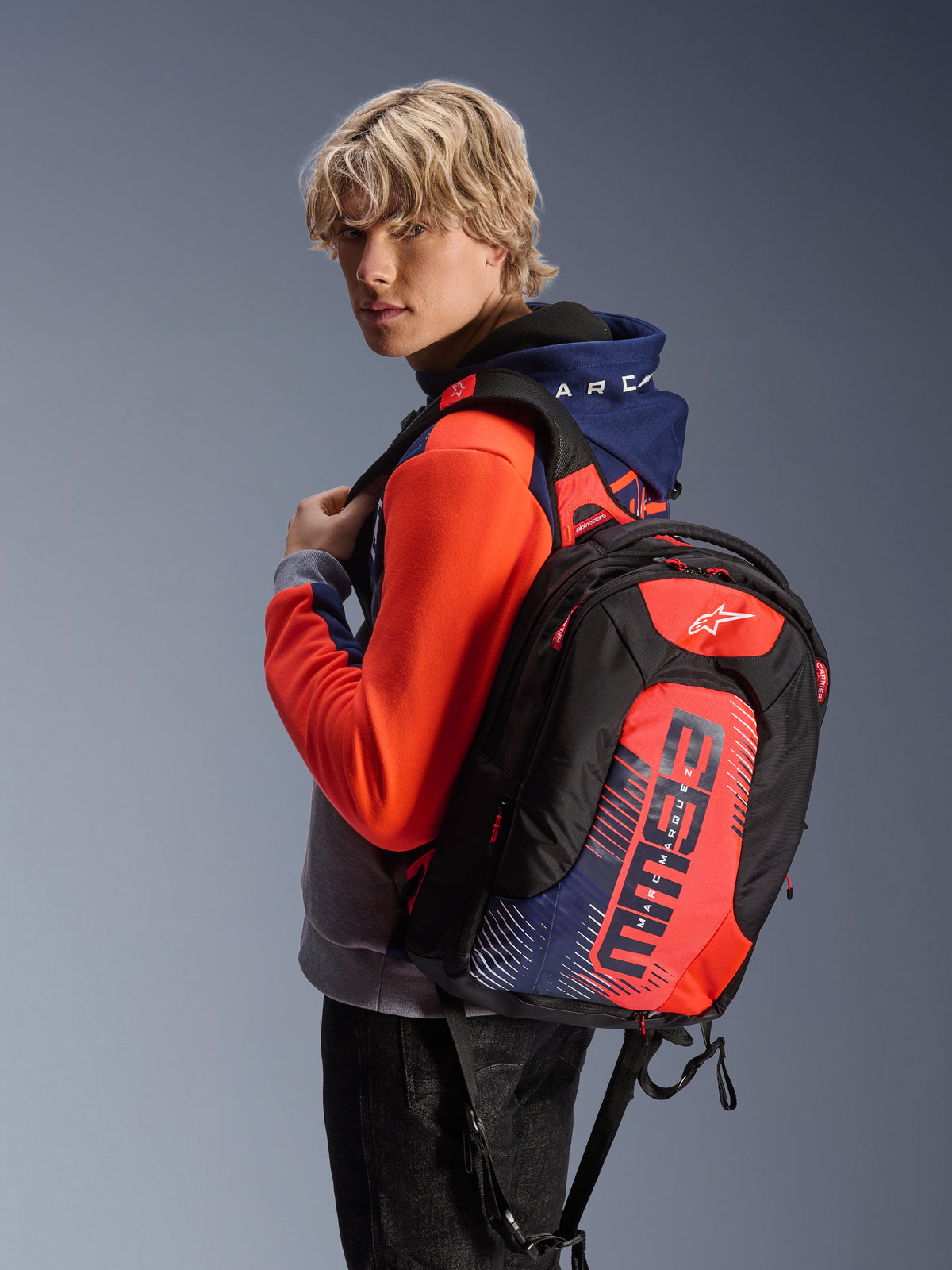 Modello che indossa la felpa con cappuccio Alpinestars MM93 Chrome V2 Sport in blu navy, grigio melange e rosso fluo, abbinata a uno zaino MM93 coordinato, abbigliamento da moto urbano casual con silhouette atletica moderna.