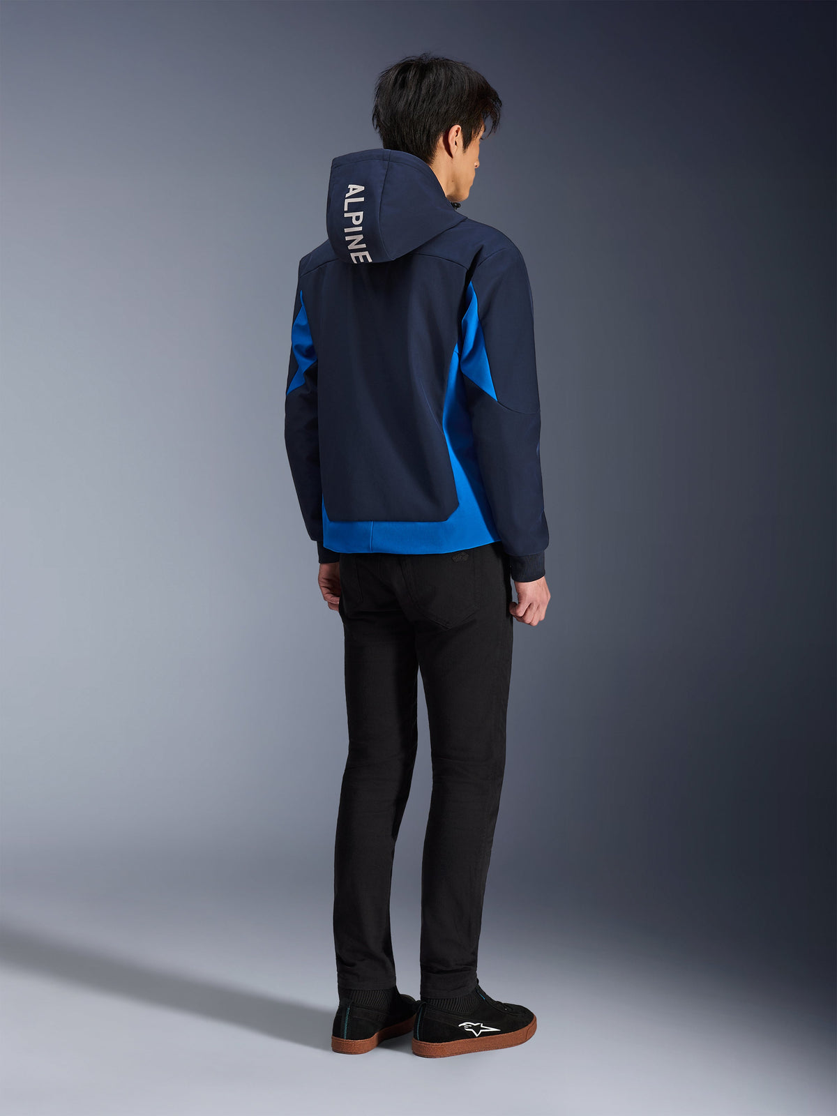Alpinestars Radium Tech Hoodie, Night Navy e Royal Blue, vista posteriore del modello che indossa la felpa tecnica con scritta 'ALPINE' bianca sul cappuccio, abbinata a pantaloni e scarpe neri, abbigliamento da moto urbano
